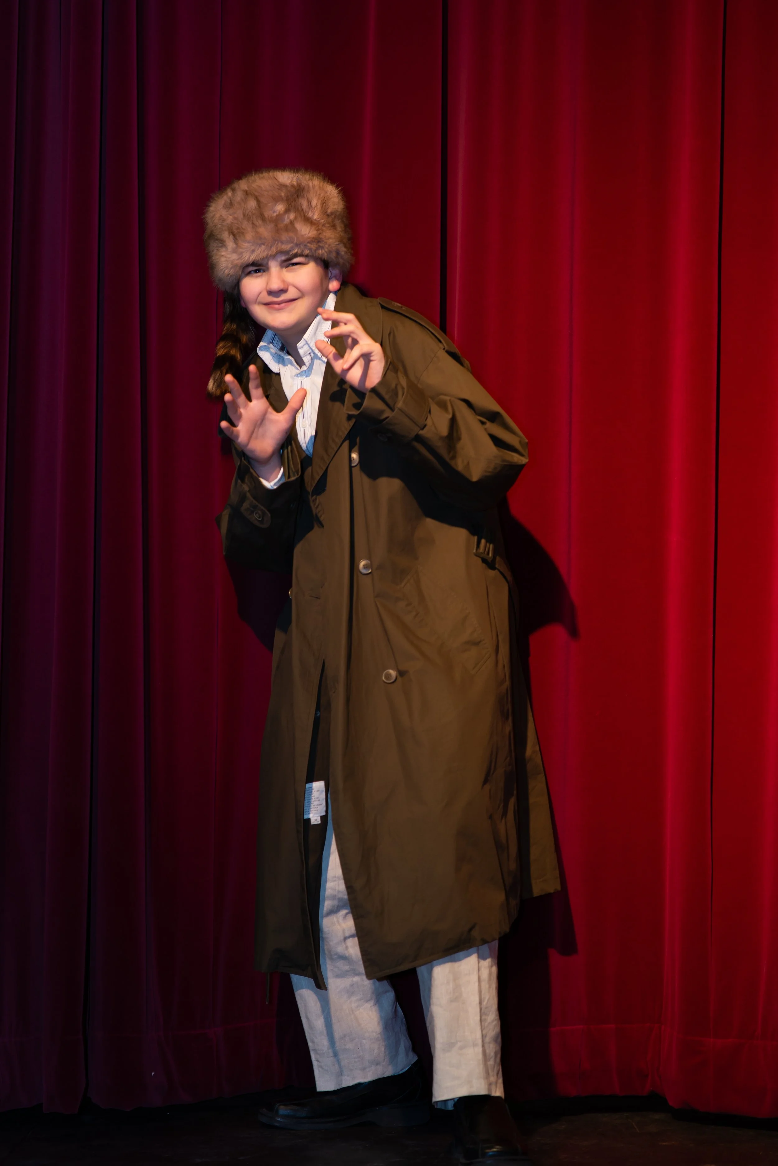 Person wearing a large fur hat and a green coat, standing in front of red curtains, making a hand gesture.