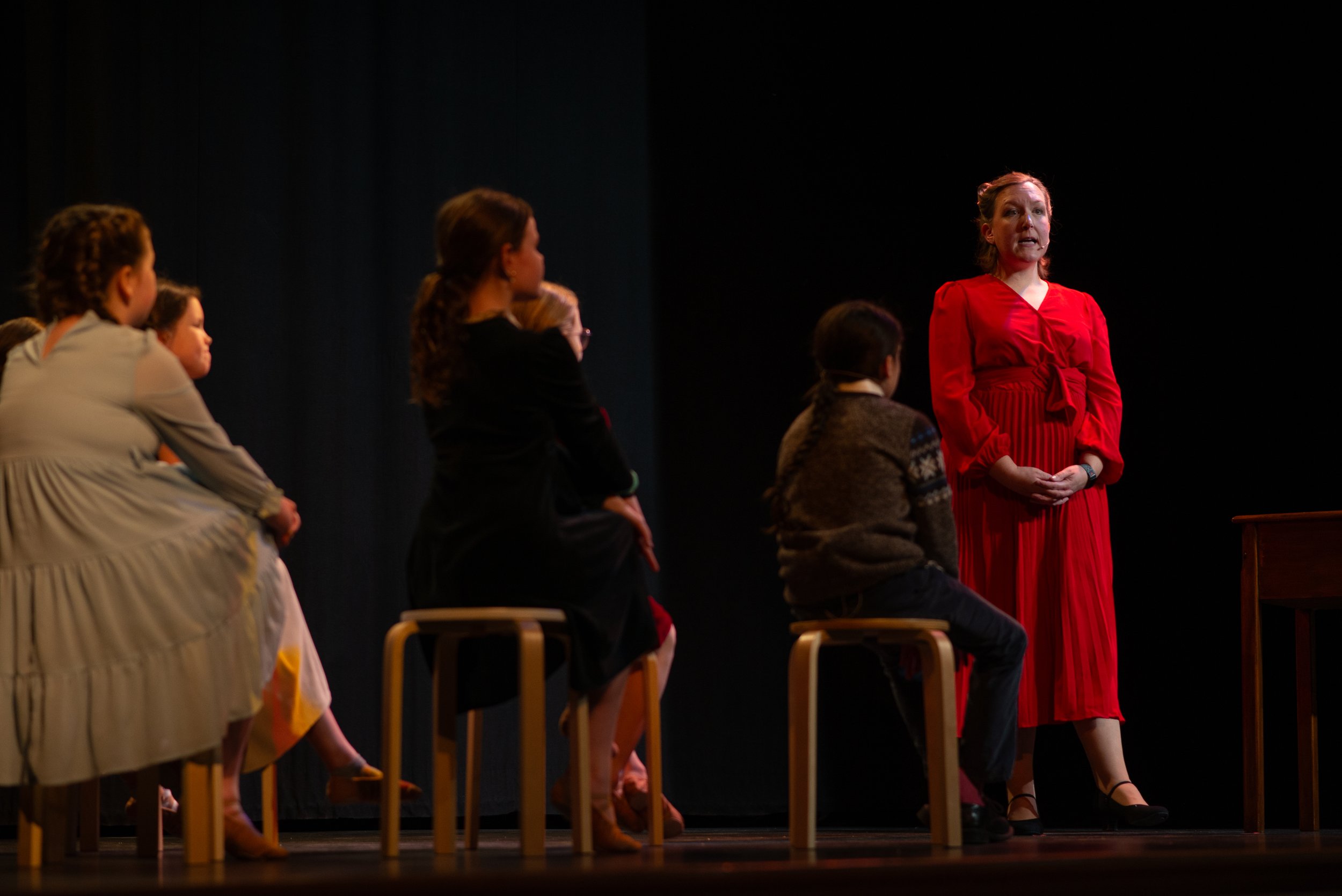 A woman in a long red dress speaking on a stage with several seated young girls watching her.