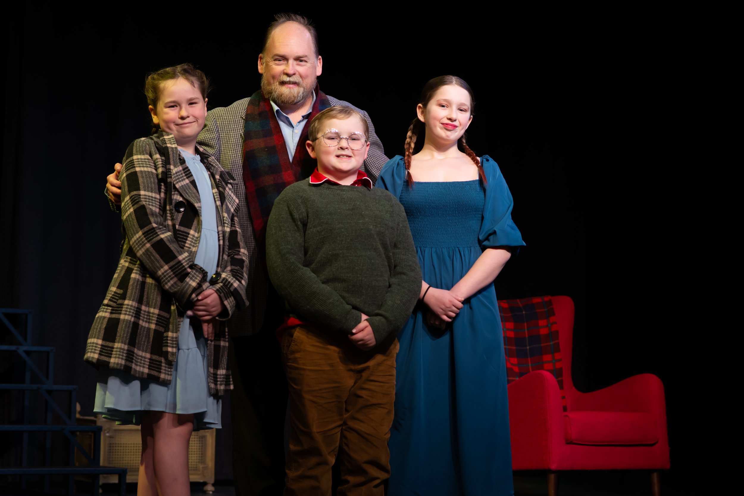 Four people, a man and three young girls, standing together on a stage with a black background. The man has a beard, glasses, and is smiling, wearing a gray coat, a shirt, and a colorful scarf. The girls have different hairstyles and outfits, and the