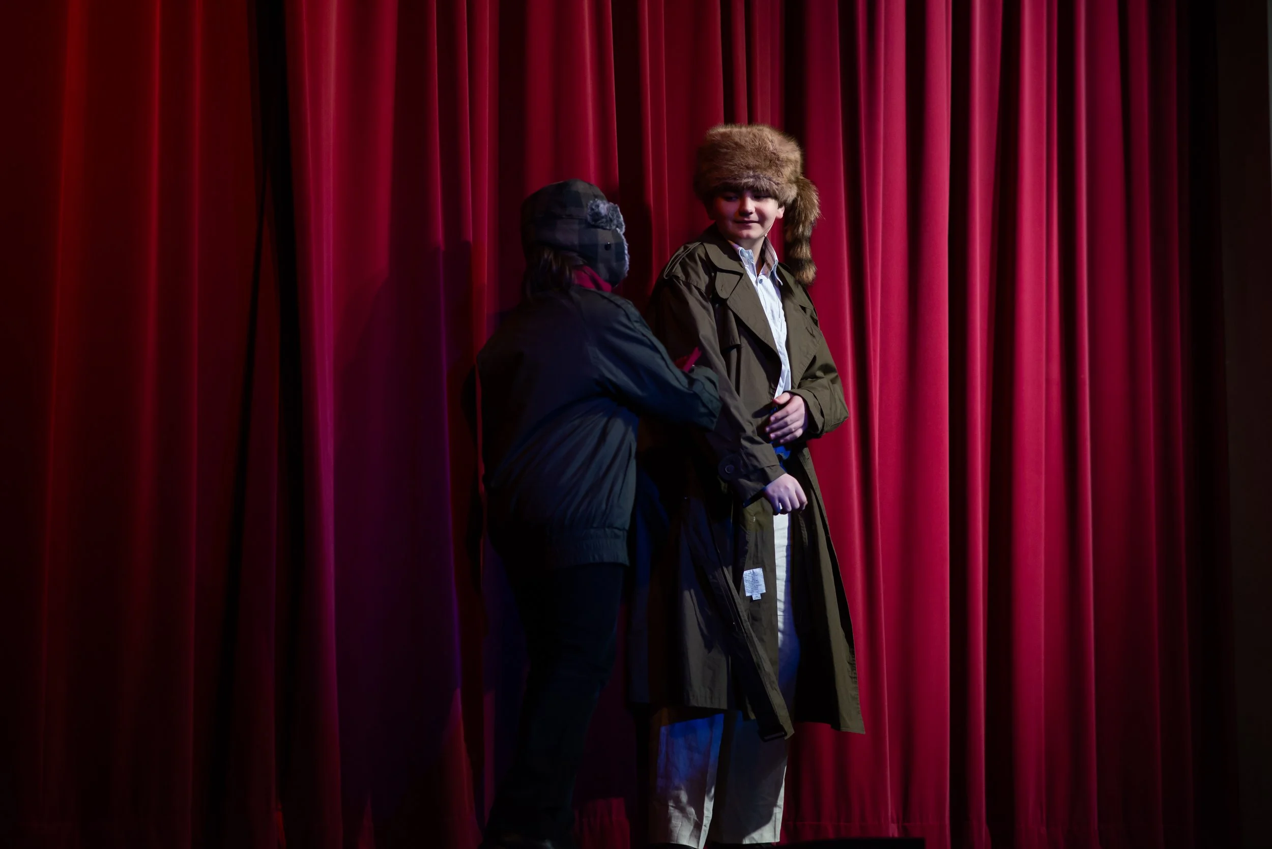 Two boys standing in front of red theater curtains, one wearing a brown coat and a fur hat, and the other wearing a dark jacket and cap.