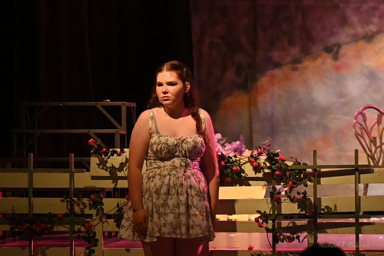 Young woman in a floral summer dress standing on stage with a backdrop of painted scenery and pink flowers in the foreground, appearing concerned or upset.