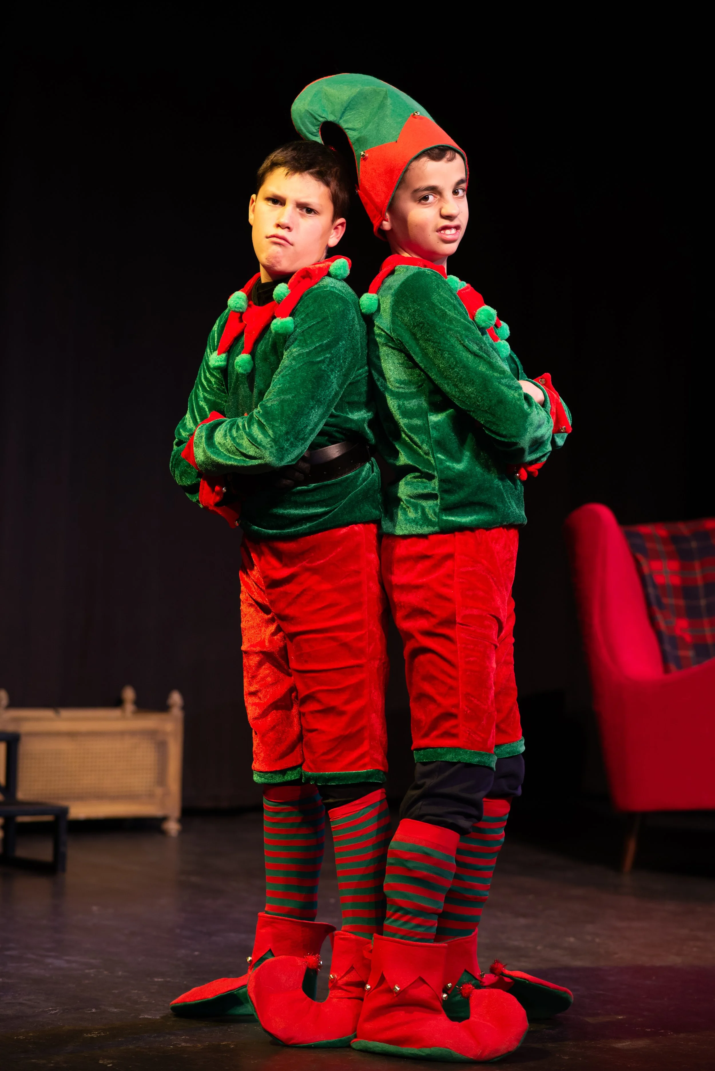 Two boys dressed as Christmas elves standing on stage with arms crossed, wearing green and red elf costumes with hats, striped stockings, and elf shoes.
