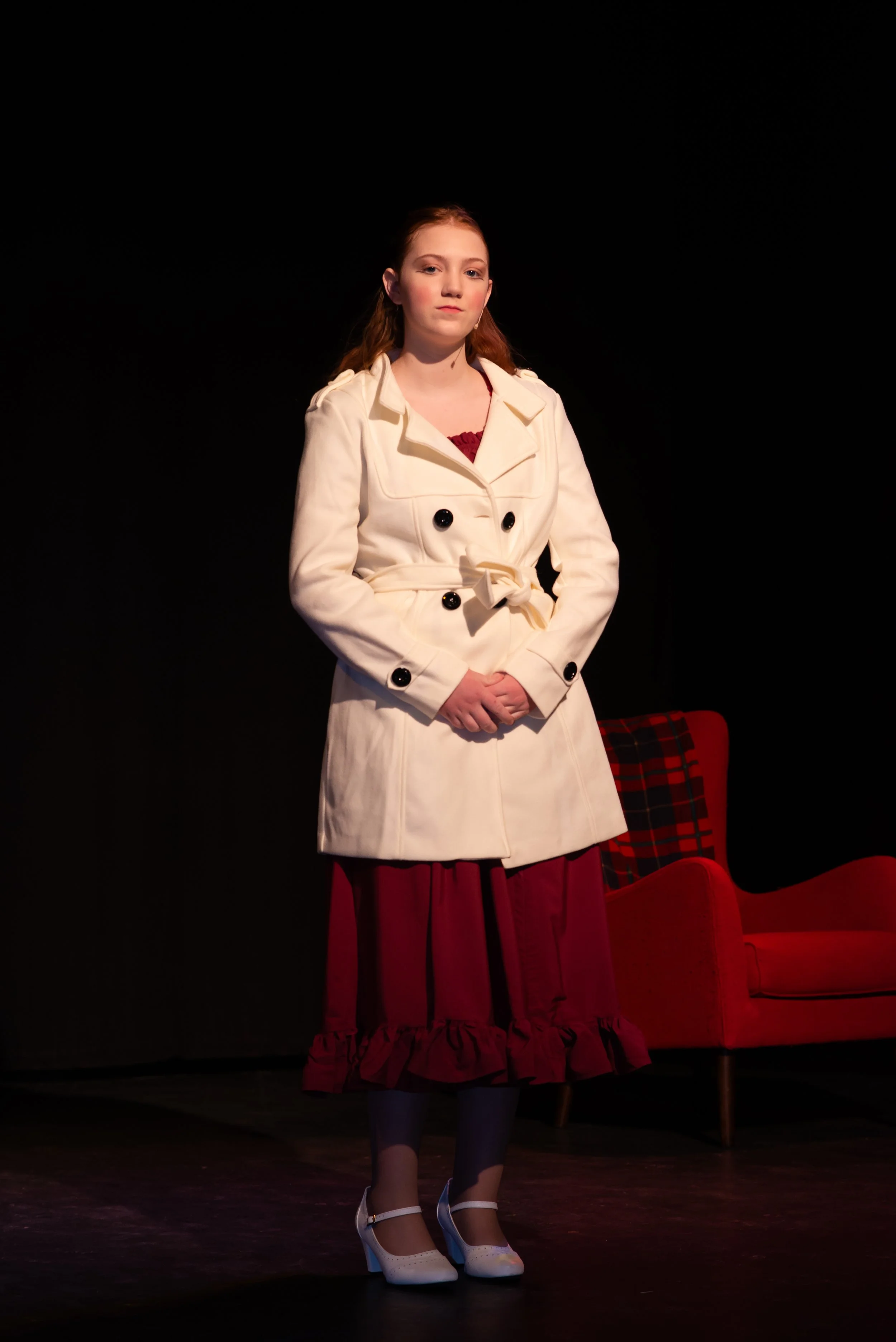A young woman with red hair stands on stage, wearing a cream-colored coat over a dark red dress, with white heels, in front of a black background with red chairs.