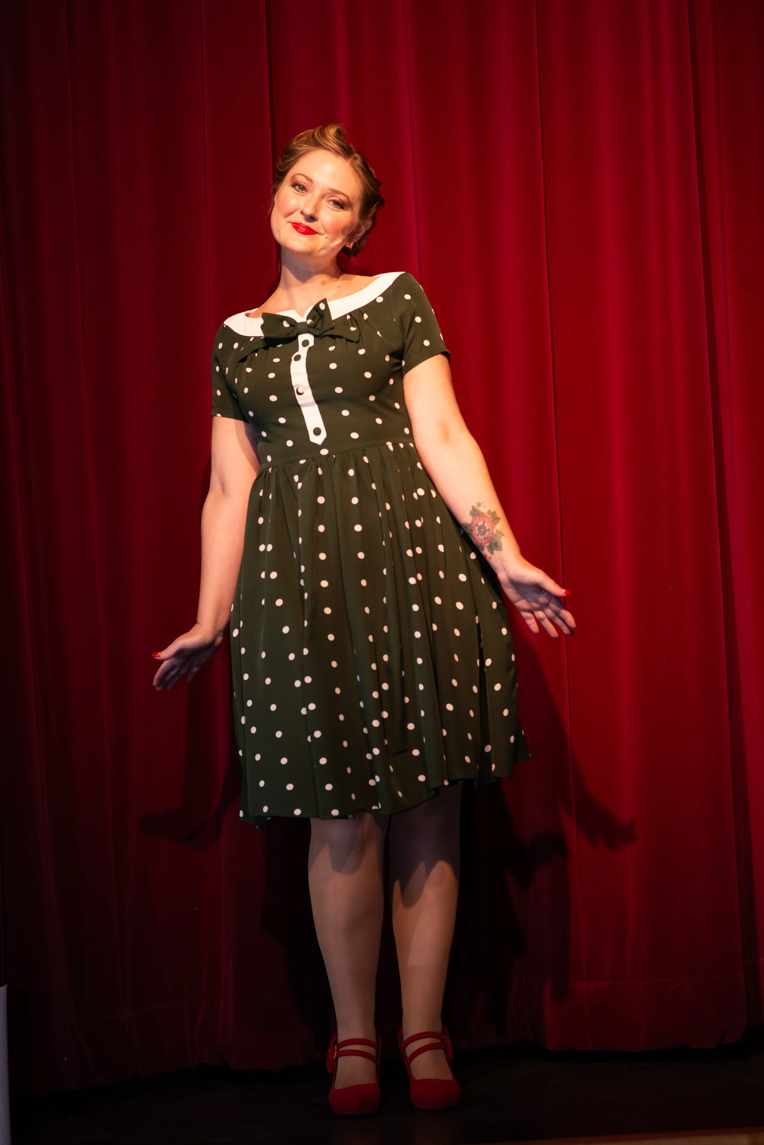 Woman in vintage black and white polka dot dress with red heels standing in front of a red curtain