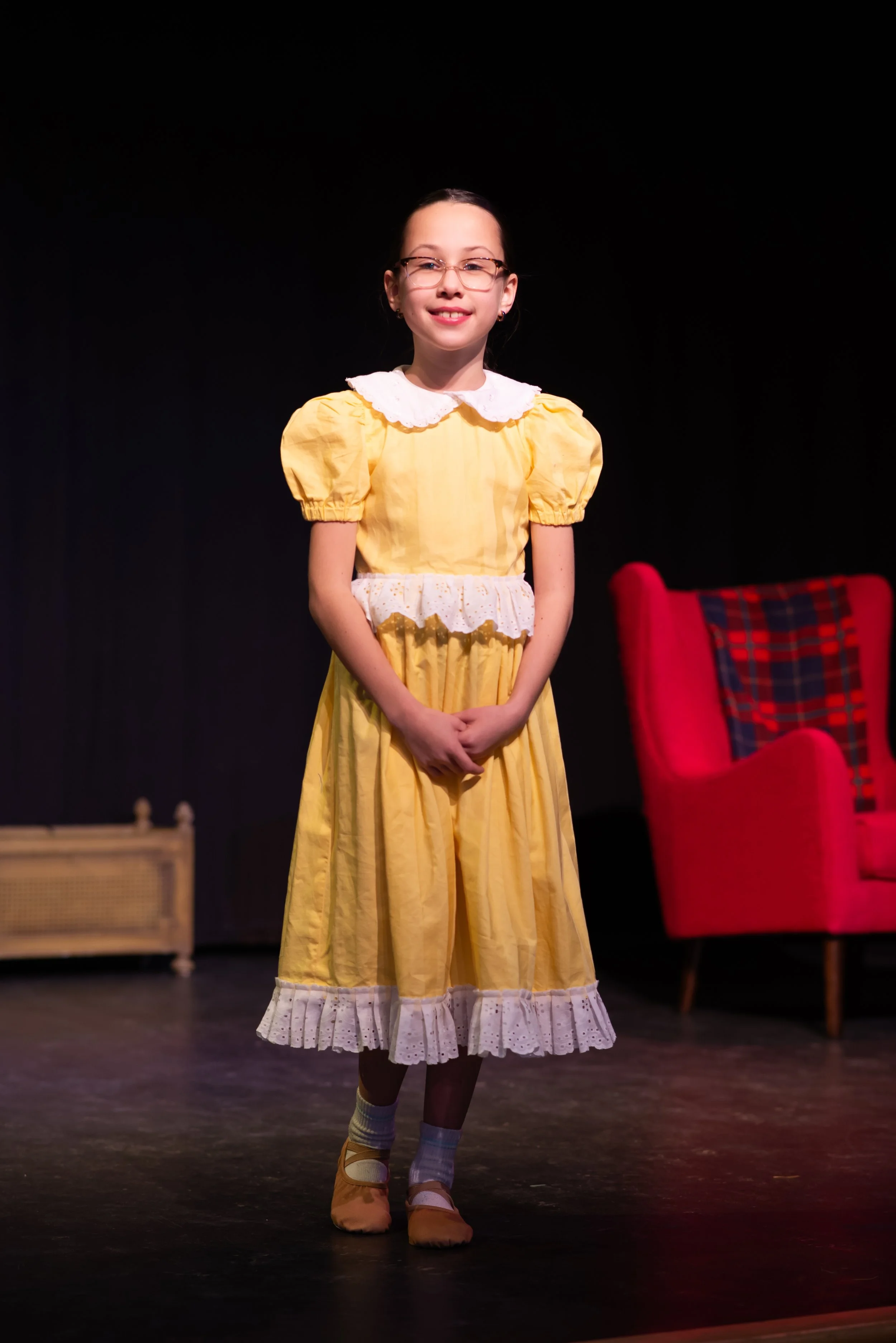 Young girl with glasses wearing a yellow dress with a white lace collar and apron, standing on a stage with a dark background, a red upholstered chair with plaid cushion is partially visible on her right.