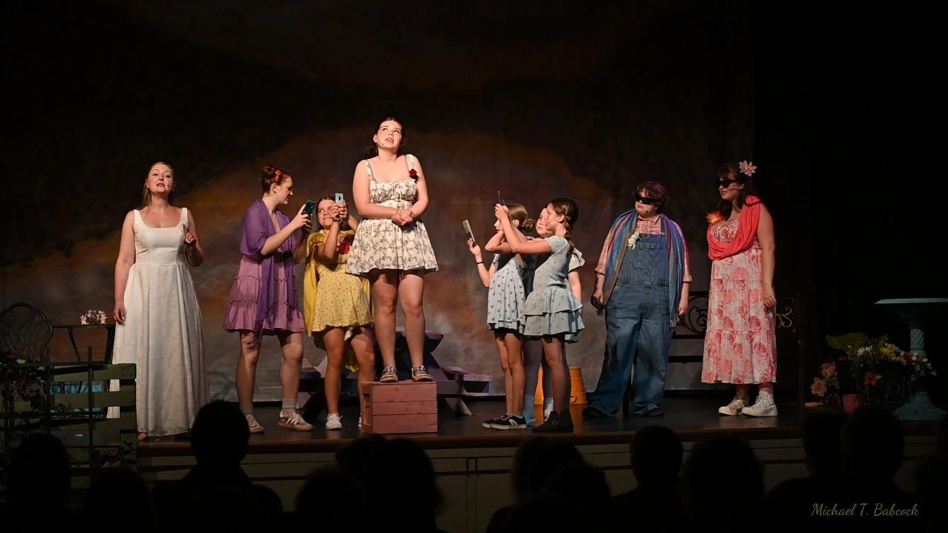 A group of women and girls performing on stage, some taking photos, in a theatrical play with a dark backdrop and stage props.