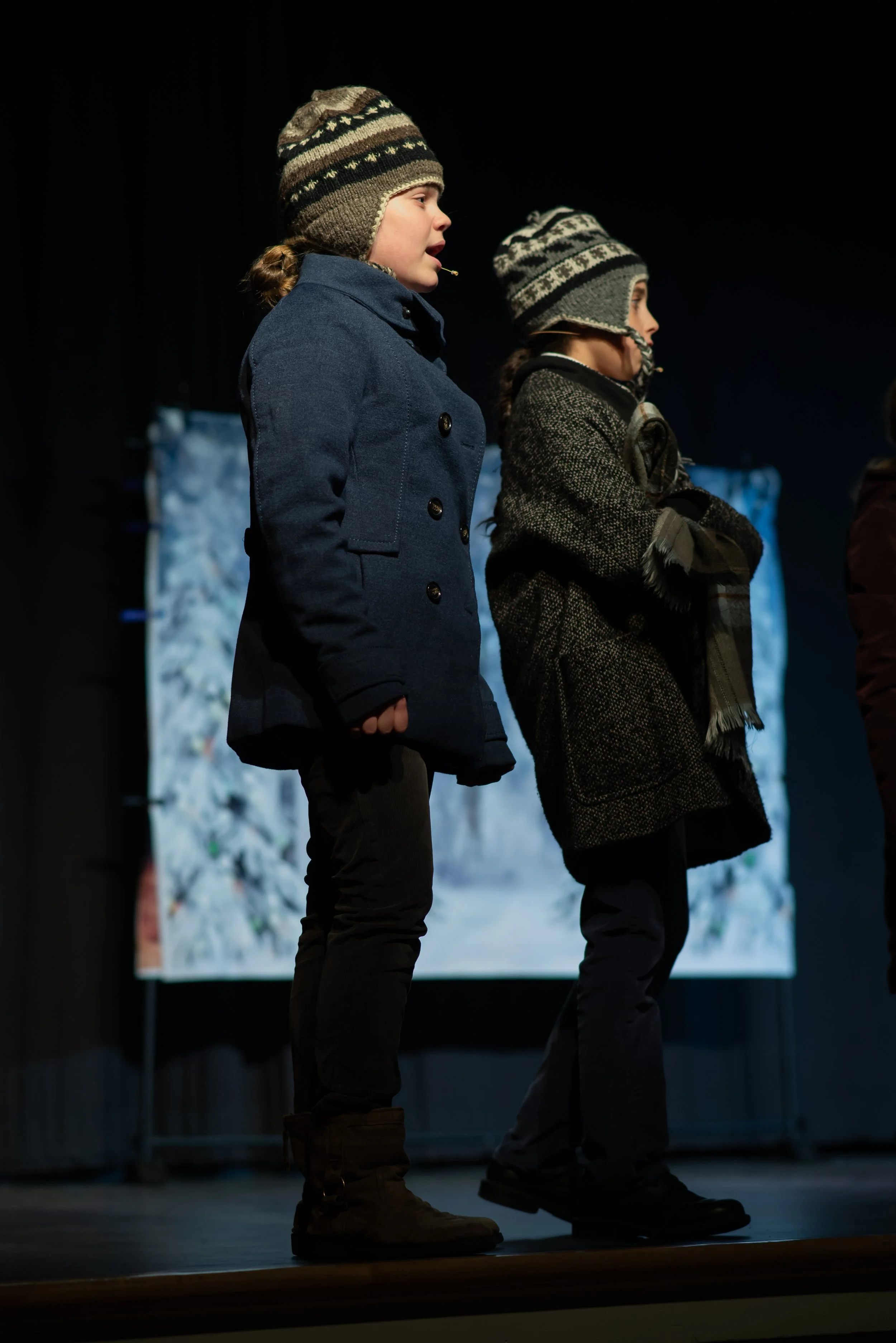Two children wearing winter coats, hats, and boots stand side by side on a stage, facing towards an audience, with a snowy backdrop behind them.