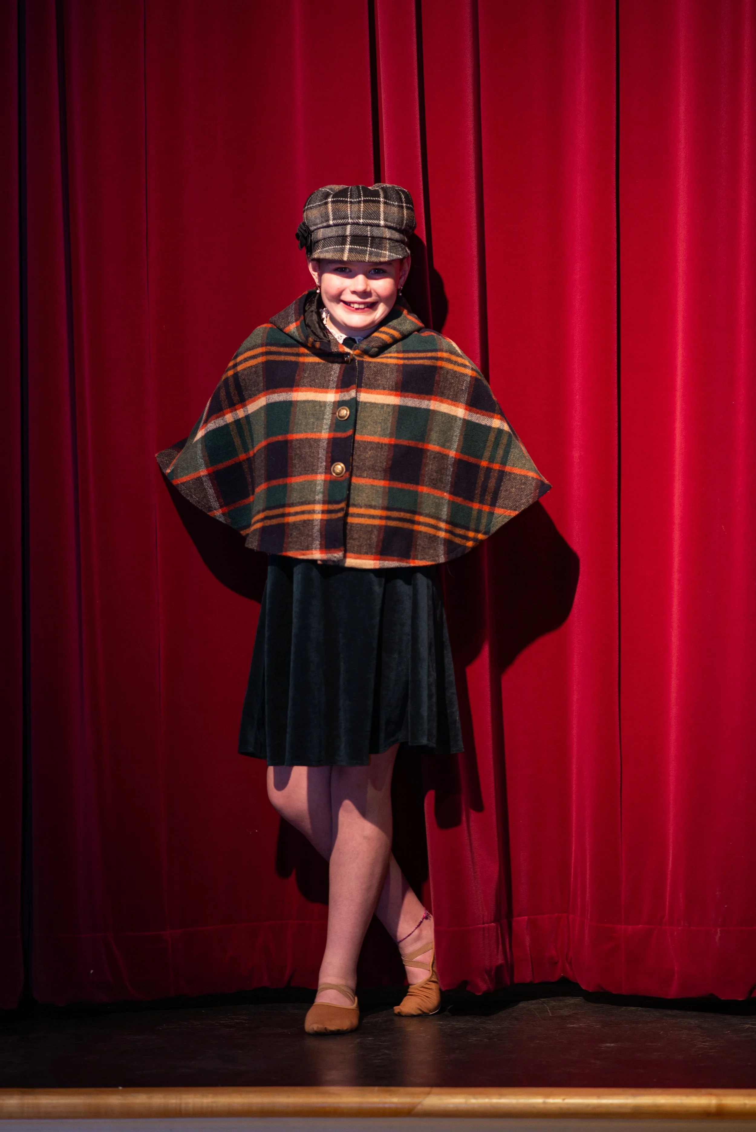 A young girl stands on stage in front of a red curtain. She is smiling and dressed in a plaid cape, a matching cap, a black skirt, and beige ballet shoes with pink ribbons.