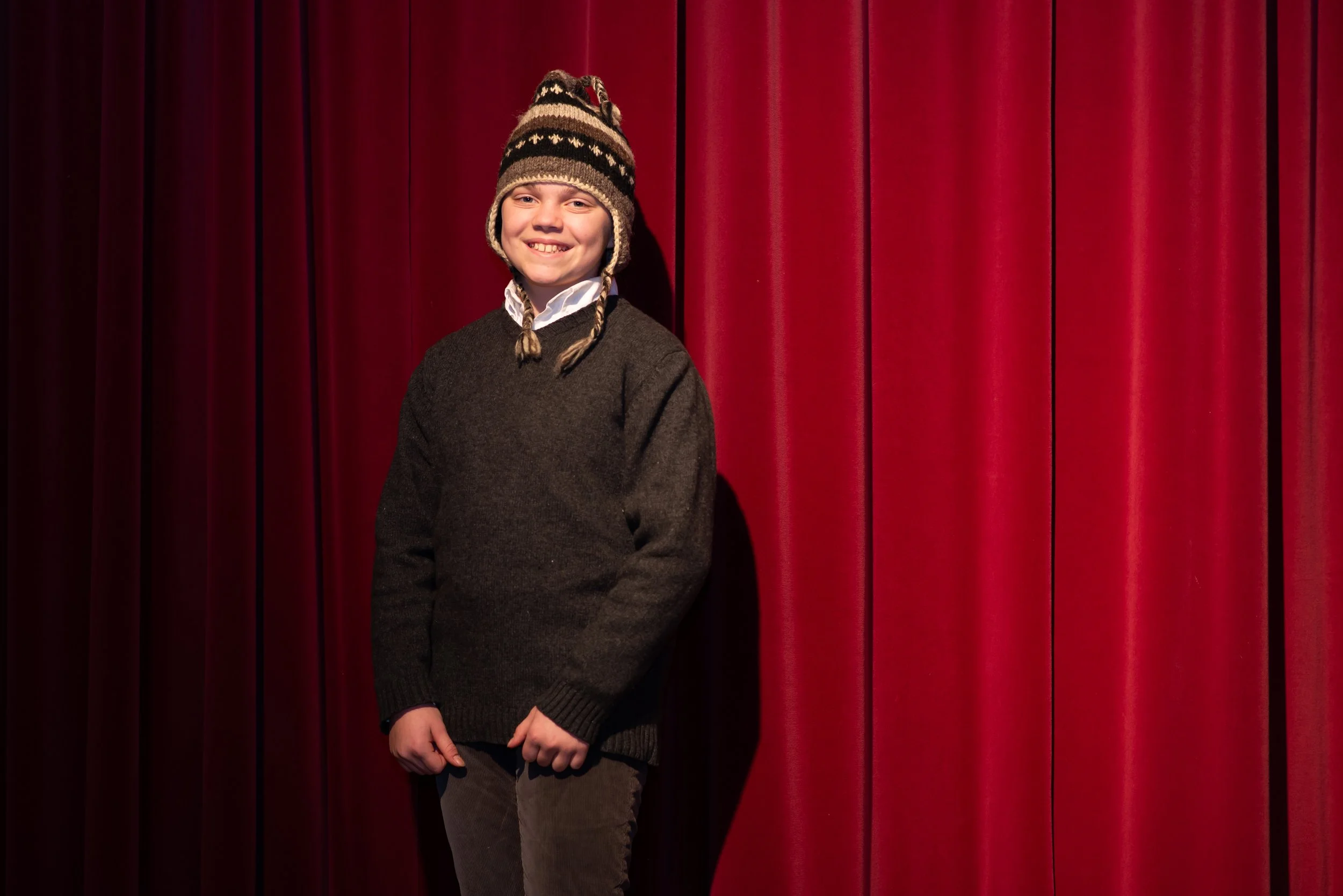 Smiling boy with a winter hat standing in front of red stage curtains.