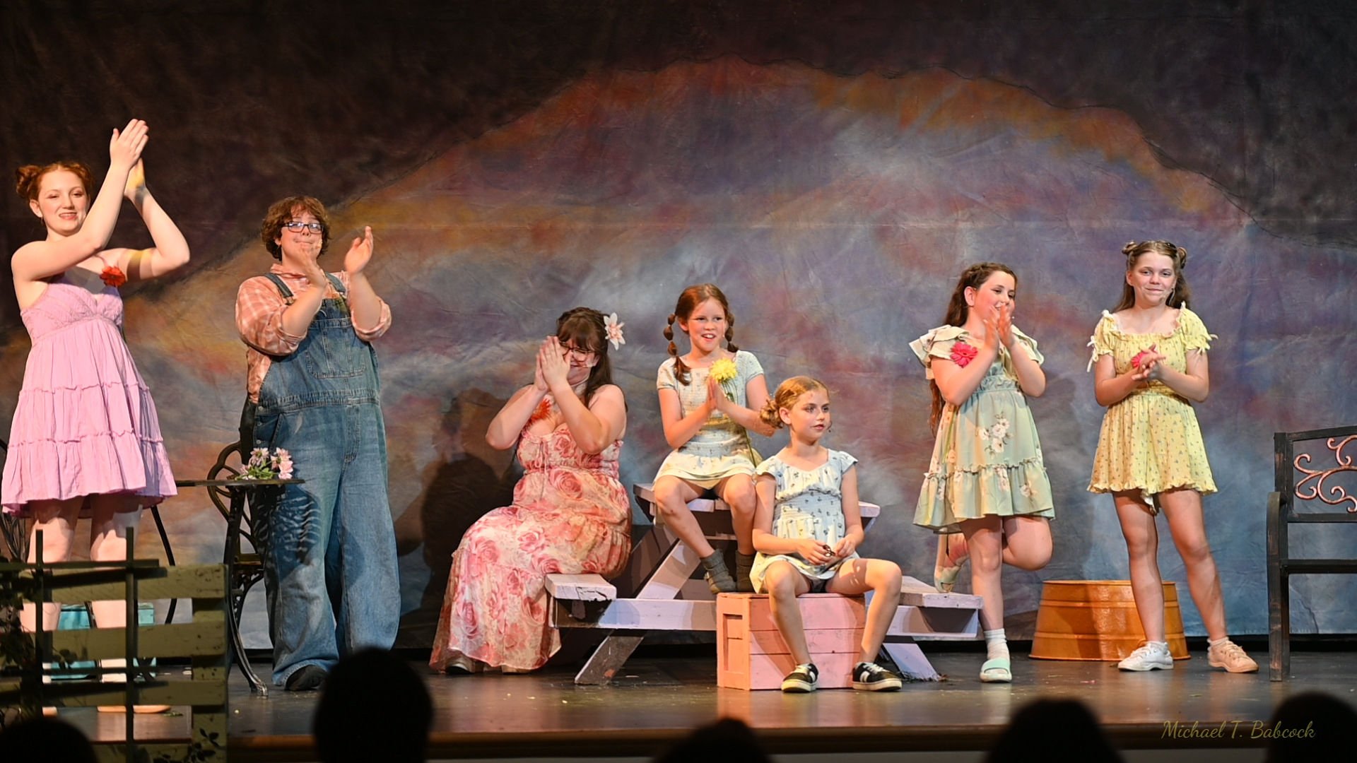 Godspell Cast standing and sitting on stage, clapping and smiling, dressed in colorful, casual summer outfits with a painted backdrop behind them.