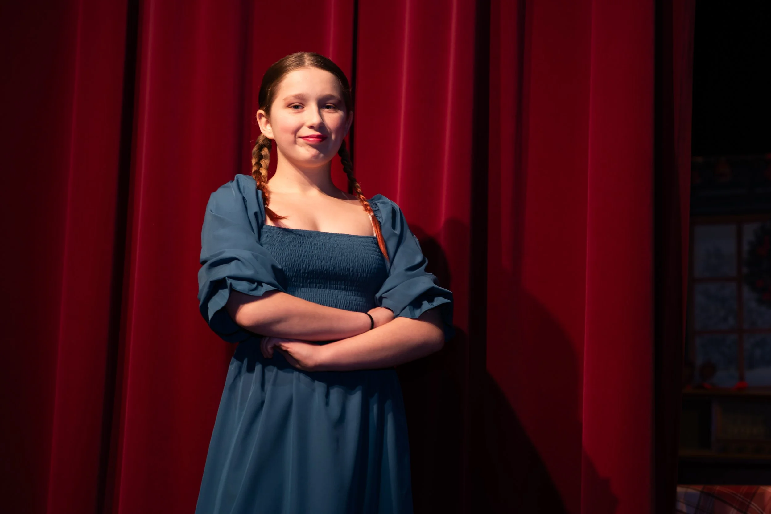 A young woman with red hair styled in two braids, wearing a blue dress with puffed sleeves, standing with arms crossed in front of red theater curtains, smiling softly.