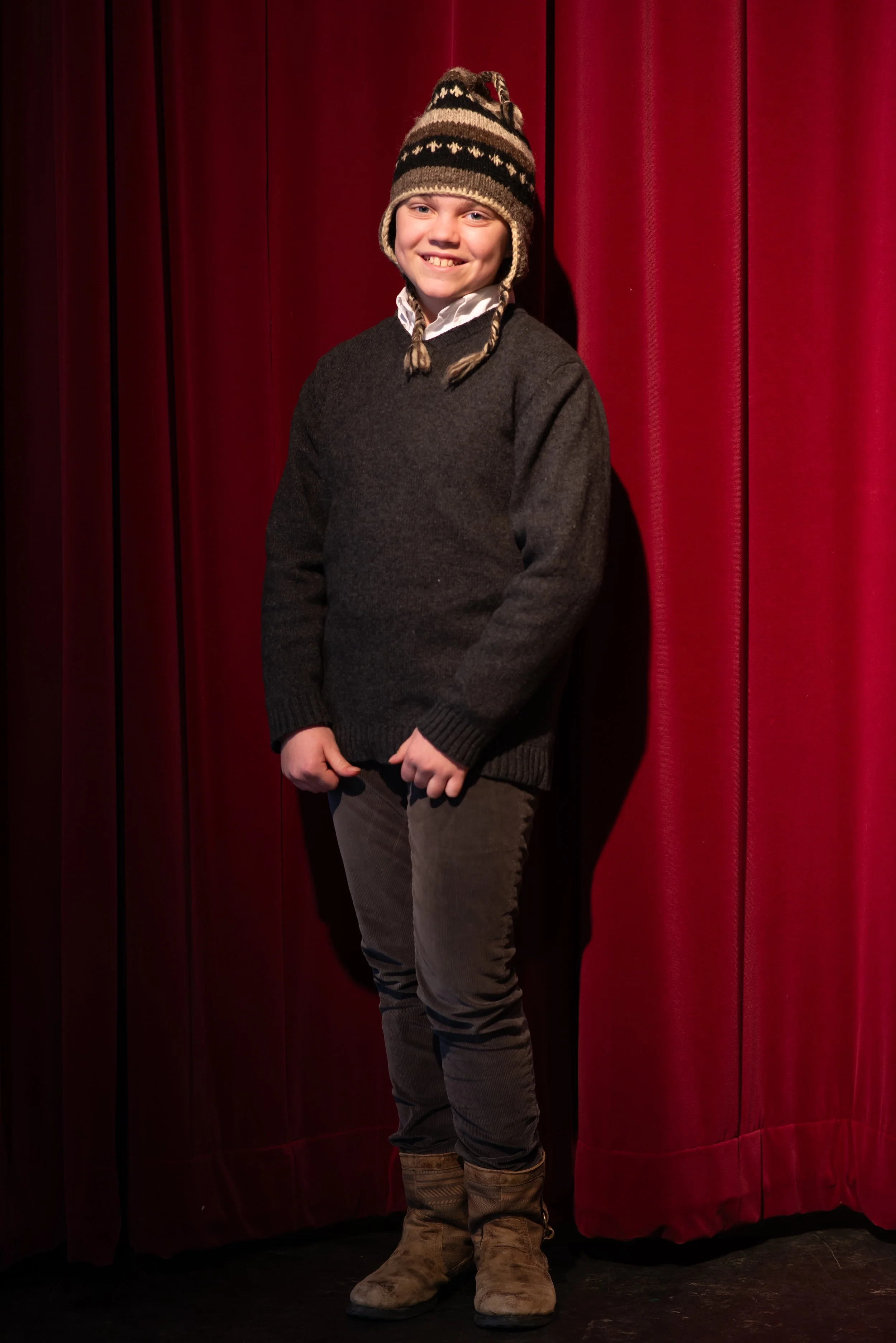 A young boy with a big smile wearing a striped knit winter hat, a dark sweater, khaki pants, and cowboy boots, standing in front of a red stage curtain.