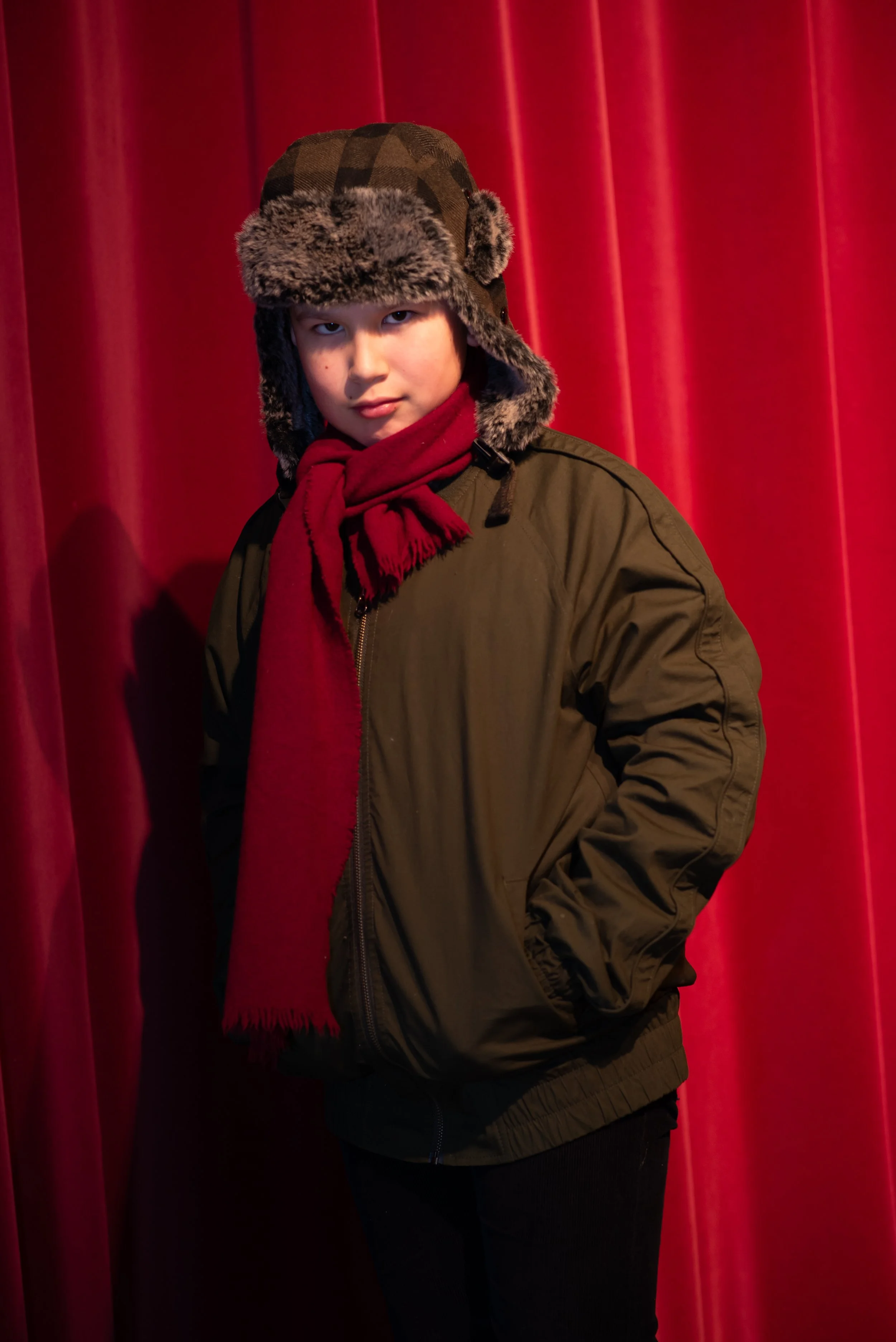 A young boy wearing a camouflage winter hat with ear flaps and faux fur trim, a red scarf, and a green winter coat, standing in front of red stage curtains.