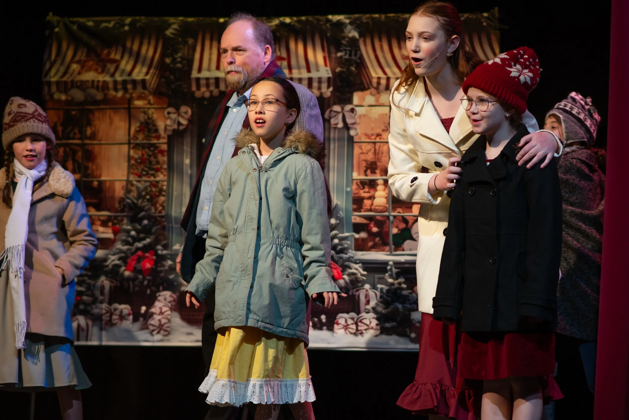 People on stage performing in a holiday-themed play, with Christmas decorations in the background including wreaths, ribbons, and decorated trees.