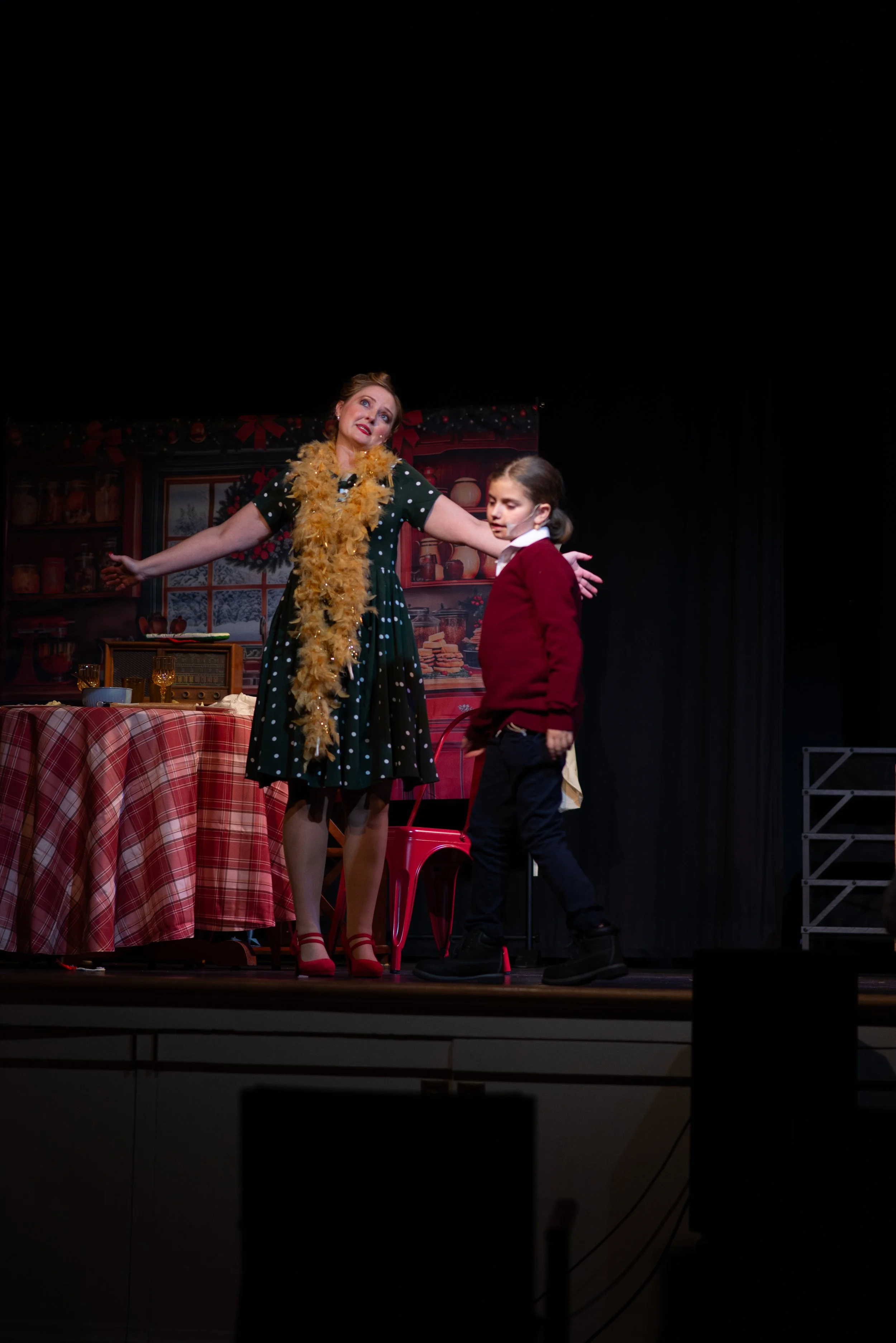 A woman dressed in a holiday-themed outfit with a yellow feather boa, speaking to a young girl wearing a red sweater on a stage with holiday decorations and a festive backdrop.