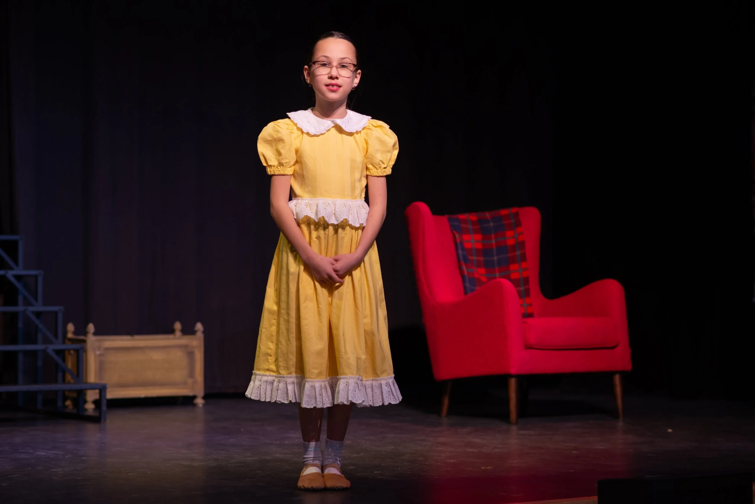Young girl standing on stage in a yellow dress with lace trim, wearing glasses, with a red and black plaid chair behind her.