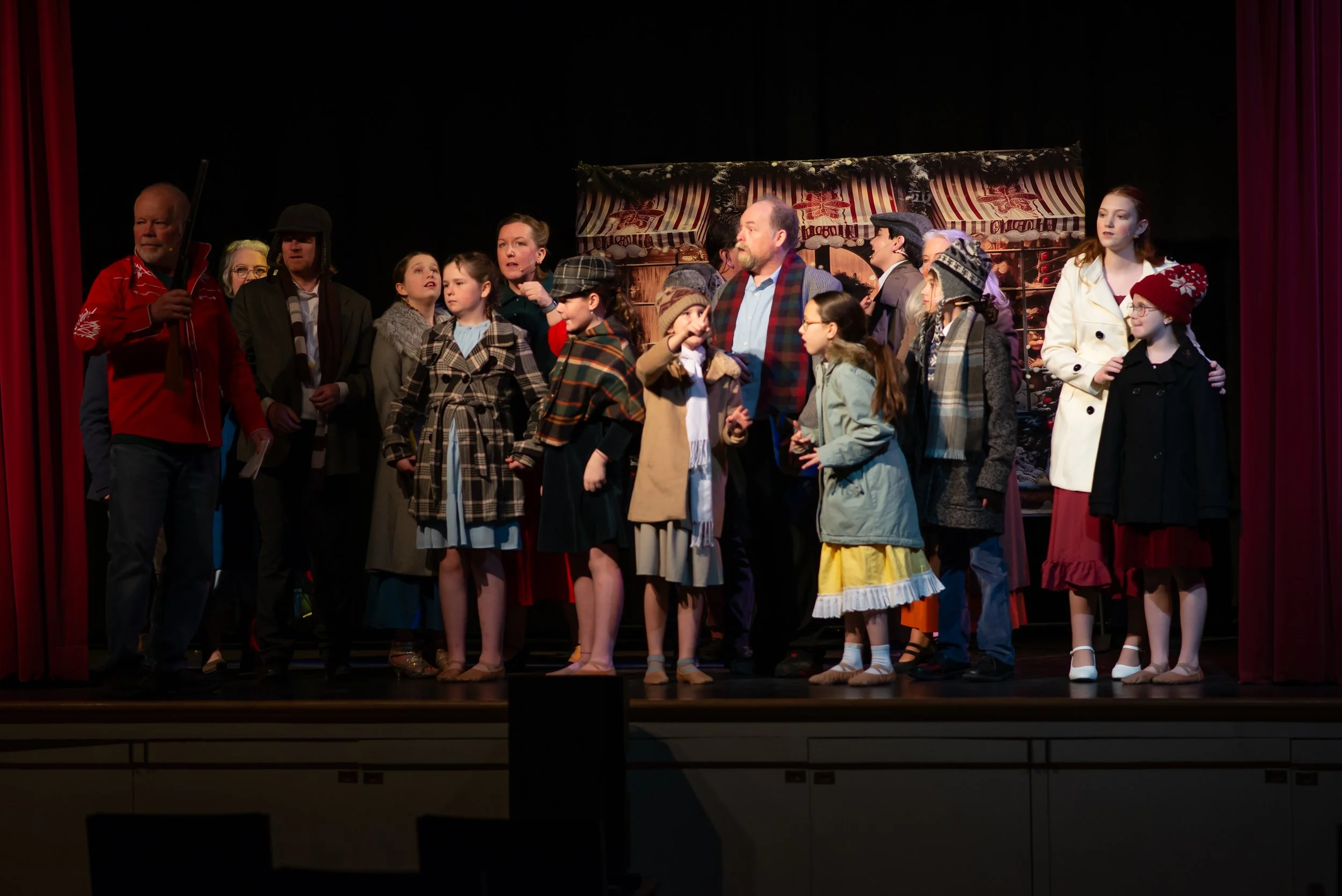 A group of actors on stage during a theatrical performance, dressed in winter-themed costumes, with a rustic Christmas set background.