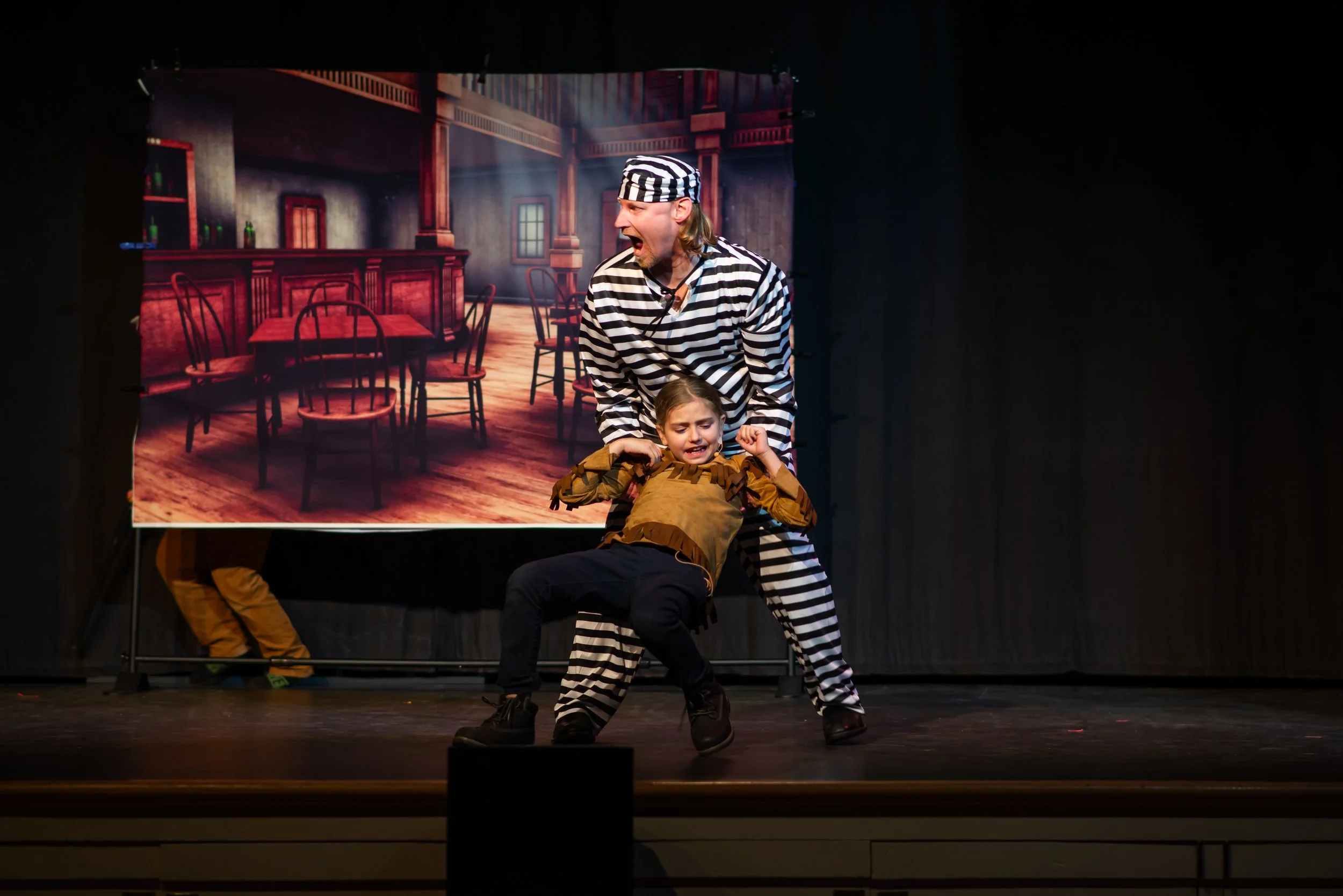 Stage performance with two actors, one dressed as a prisoner with a striped uniform and hat, gripping a young girl who appears distressed, with a backdrop of a rustic wooden room.