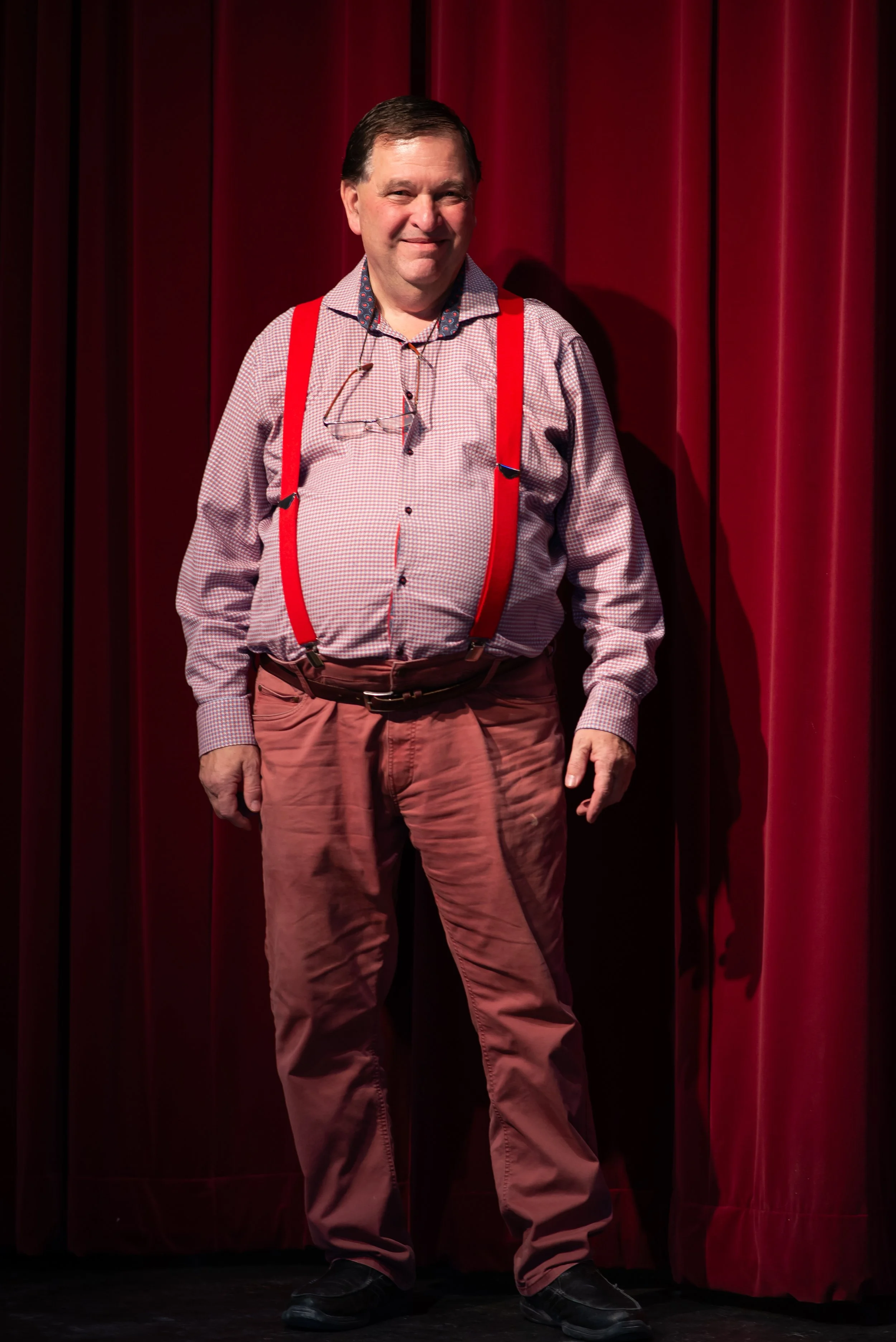 A man standing in front of red curtains, smiling, wearing a checkered shirt with a collar, red suspenders, beige pants, and black shoes. He has glasses hanging around his neck.