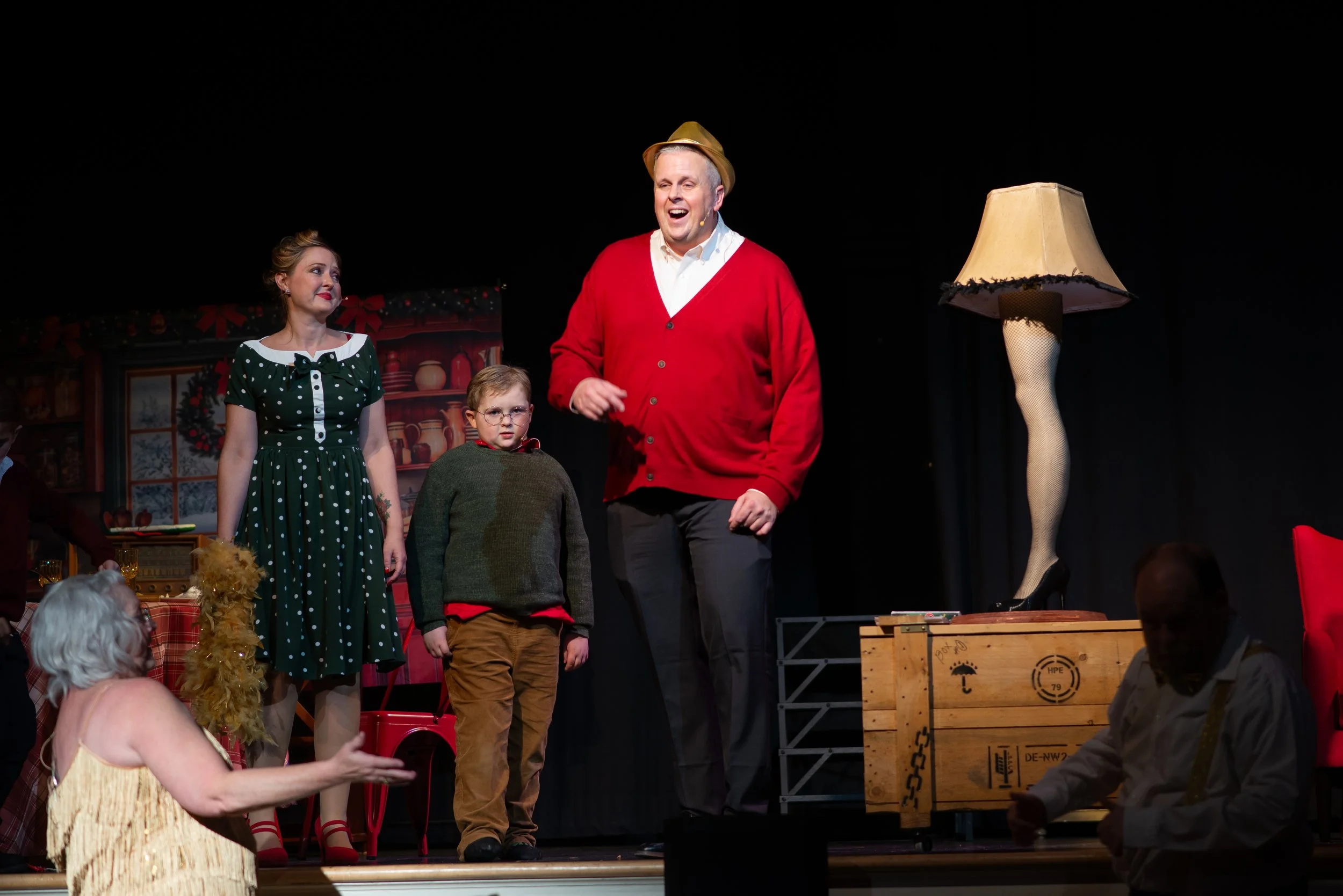 Actors on stage during a theatrical performance with Christmas decorations, including a woman in a green polka dot dress, a boy in brown and red, and a man in a red cardigan and hat, with audience members in the foreground and a lamp on the stage.