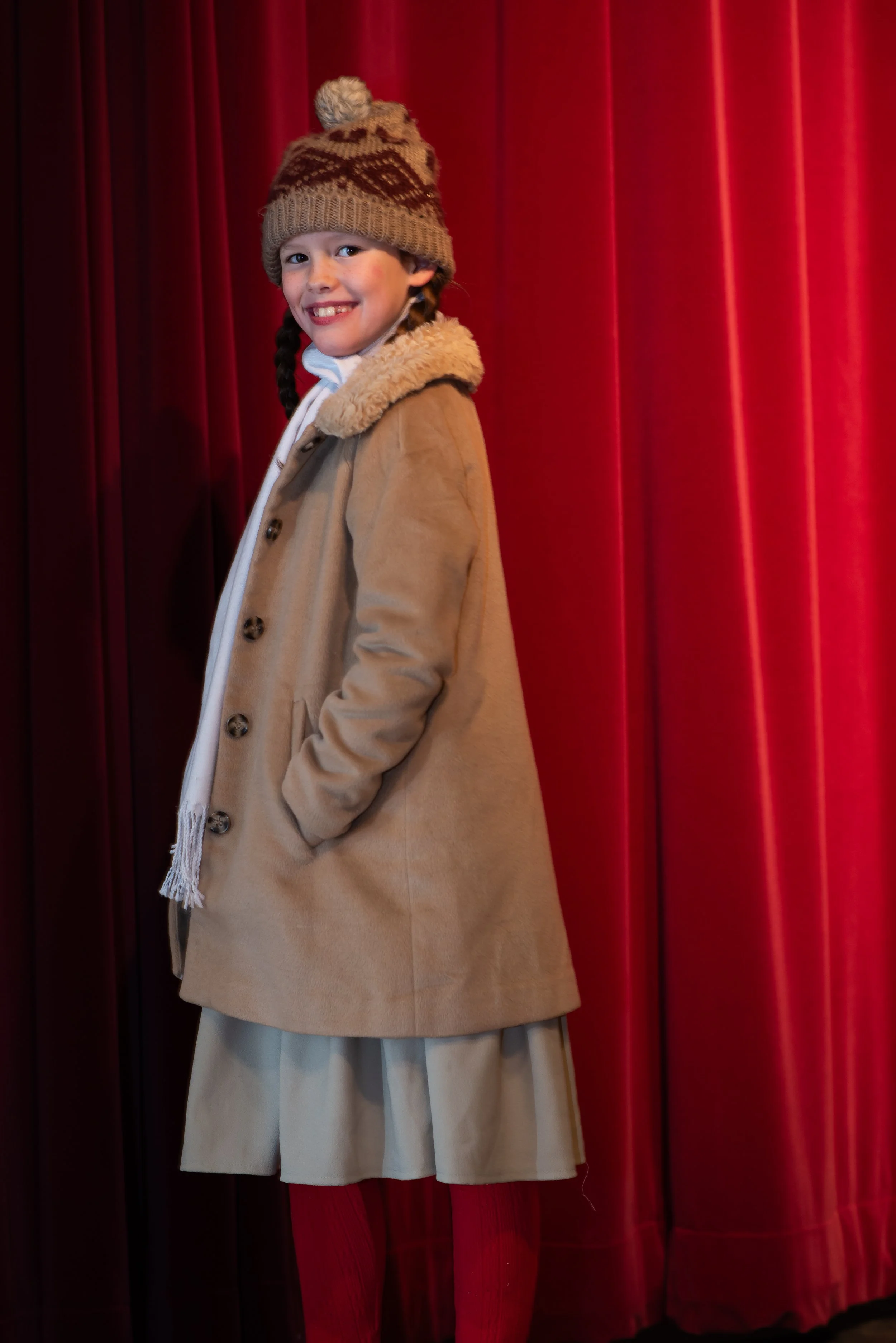 A young girl with braided hair smiling, standing in front of red curtain. She is wearing a tan coat with a furry collar, a beige and brown knit hat with a pom-pom, and beige pants.