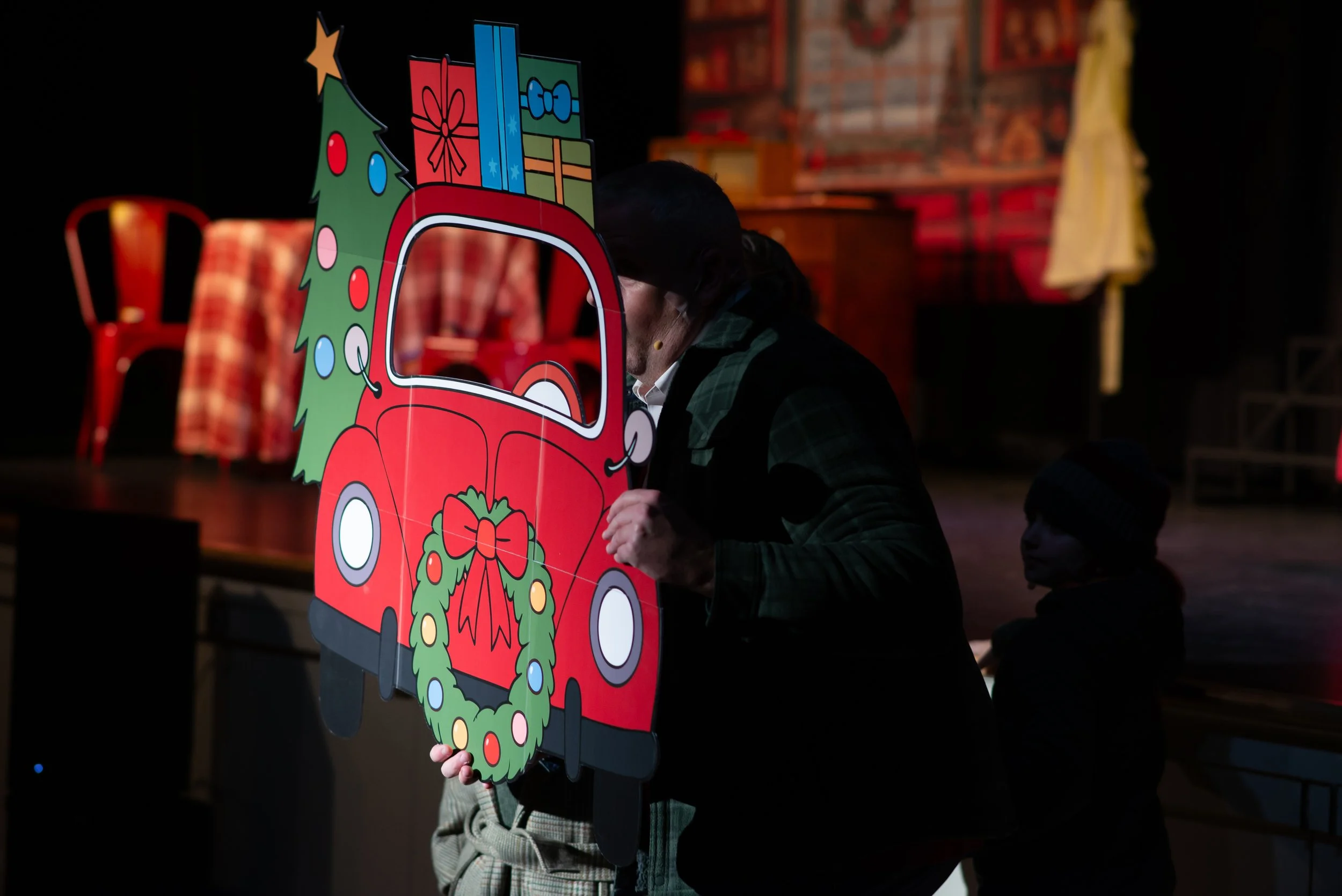 A person holding a colorful Christmas-themed cutout featuring a red car with a wreath and presents on its roof, with a Christmas tree and stars. Someone is sitting behind it, partially visible.