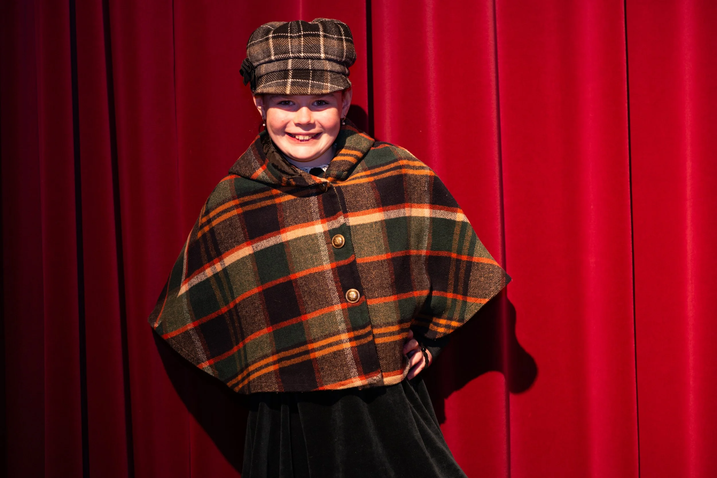 A young girl with a big smile, wearing a plaid hat and a plaid cape, standing in front of a red curtain.