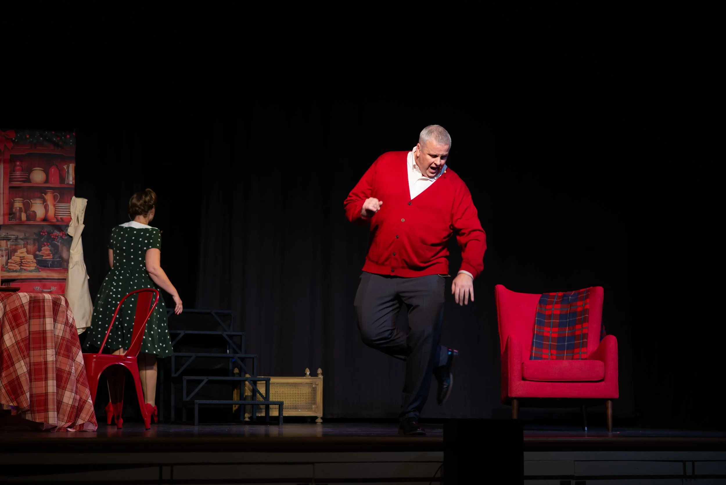 Actors performing a theatrical play on stage with Christmas-themed props and furniture, including a red armchair and a decorated backdrop.