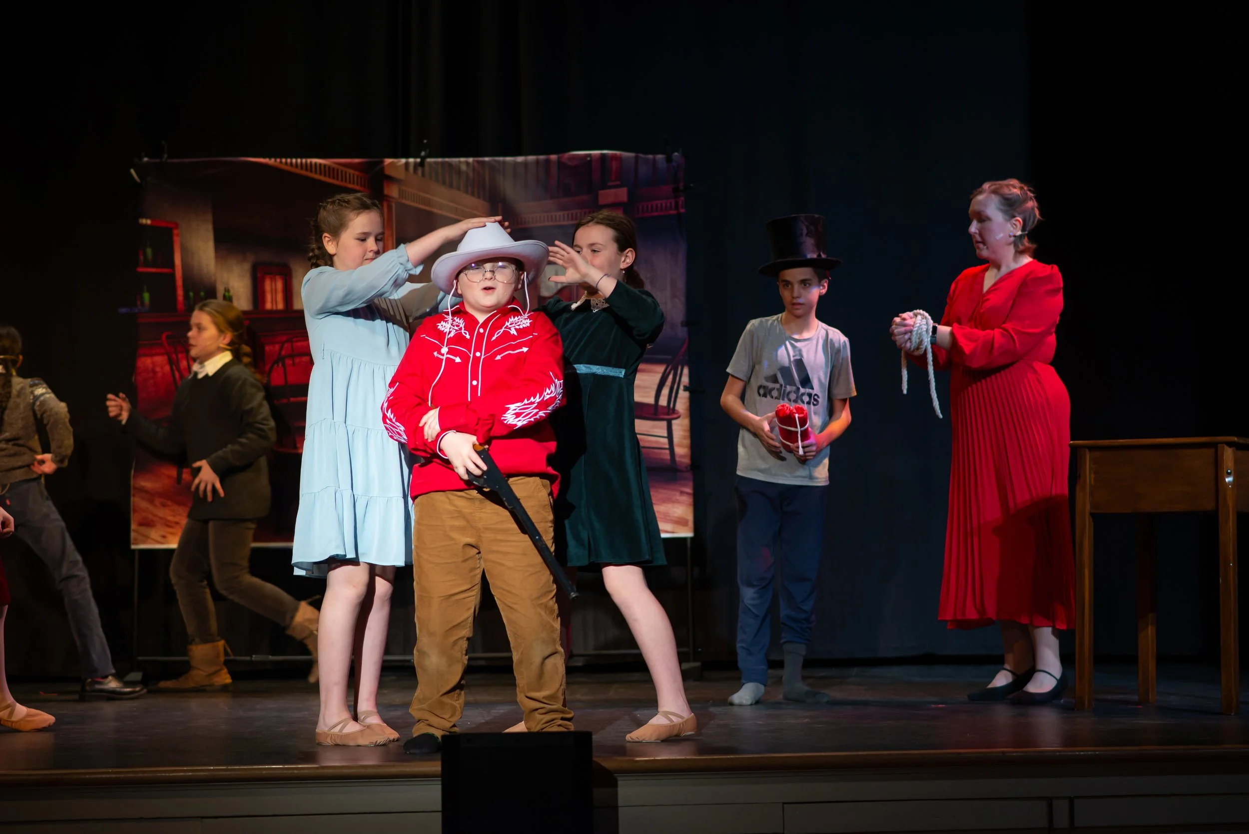 Children performing in a theatrical play, with one child wearing a cowboy hat and glasses, surrounded by other children in costumes, on a stage with a painted backdrop.