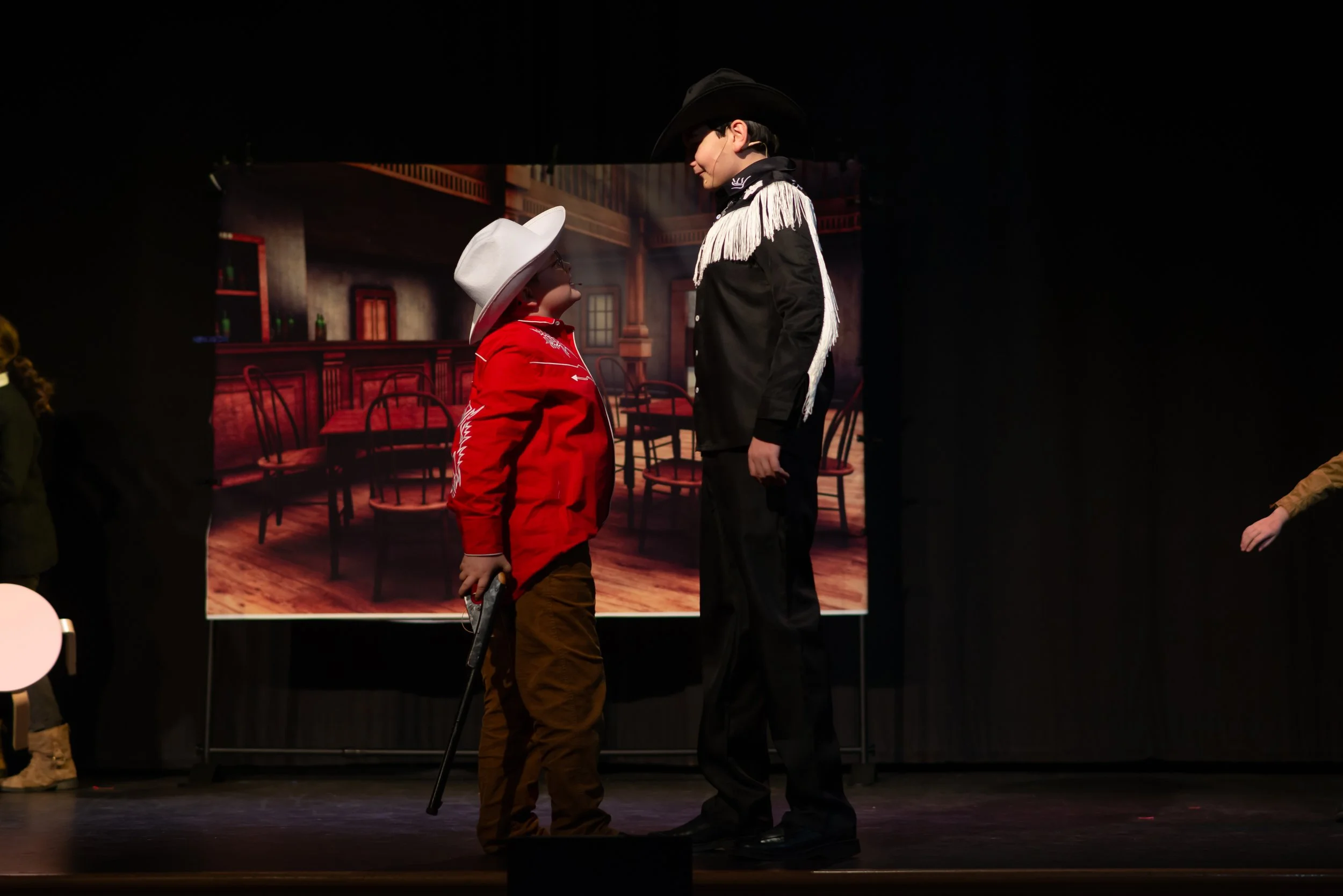 Two performers dressed as cowboys in a theatrical setting, standing face to face, with a painted backdrop of a western-style saloon interior.