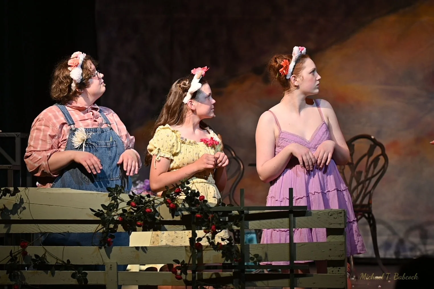 Three young women dressed in costumes on stage, looking to their right, with a garden fence with red berries and two vintage chairs in the background.