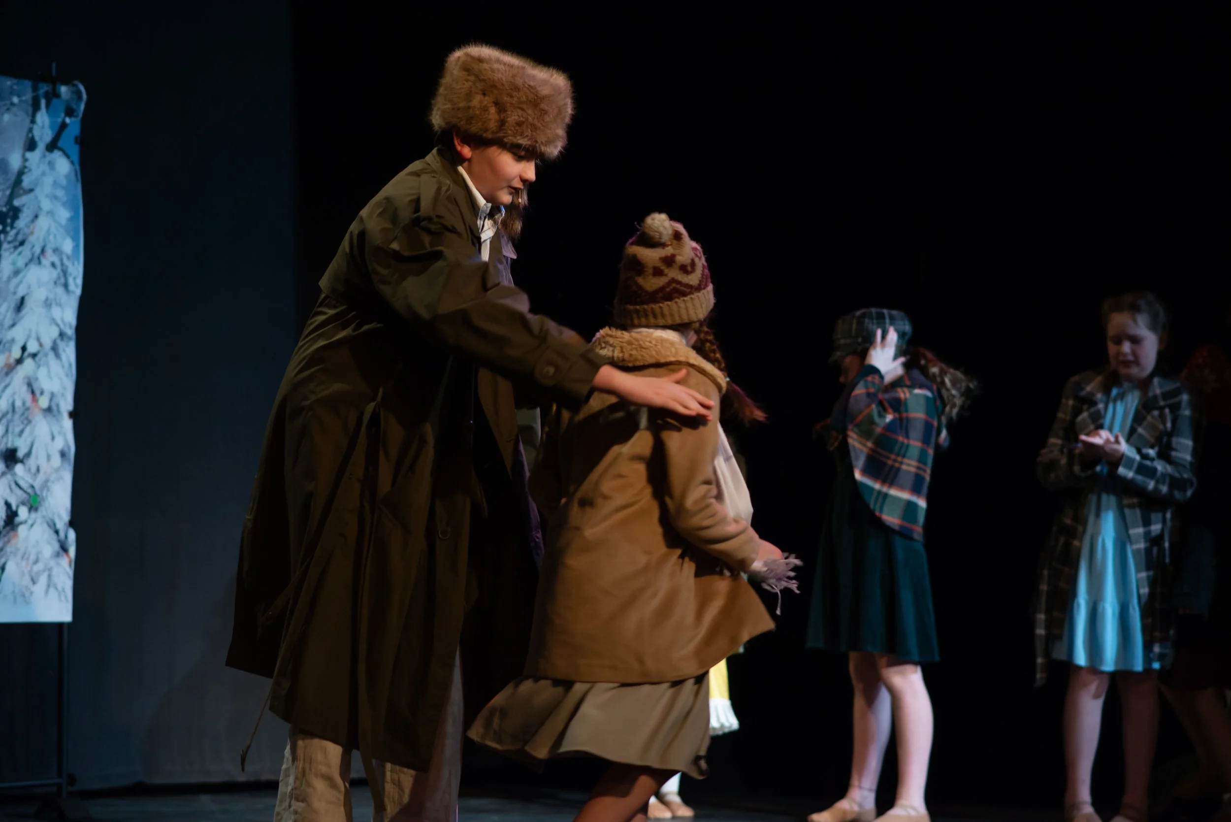 Group of children on stage, wearing winter clothing, with one adult woman in an outdoor jacket and furry hat interacting with a girl in a brown coat and knitted hat, while other children in plaid jackets and dresses look on.