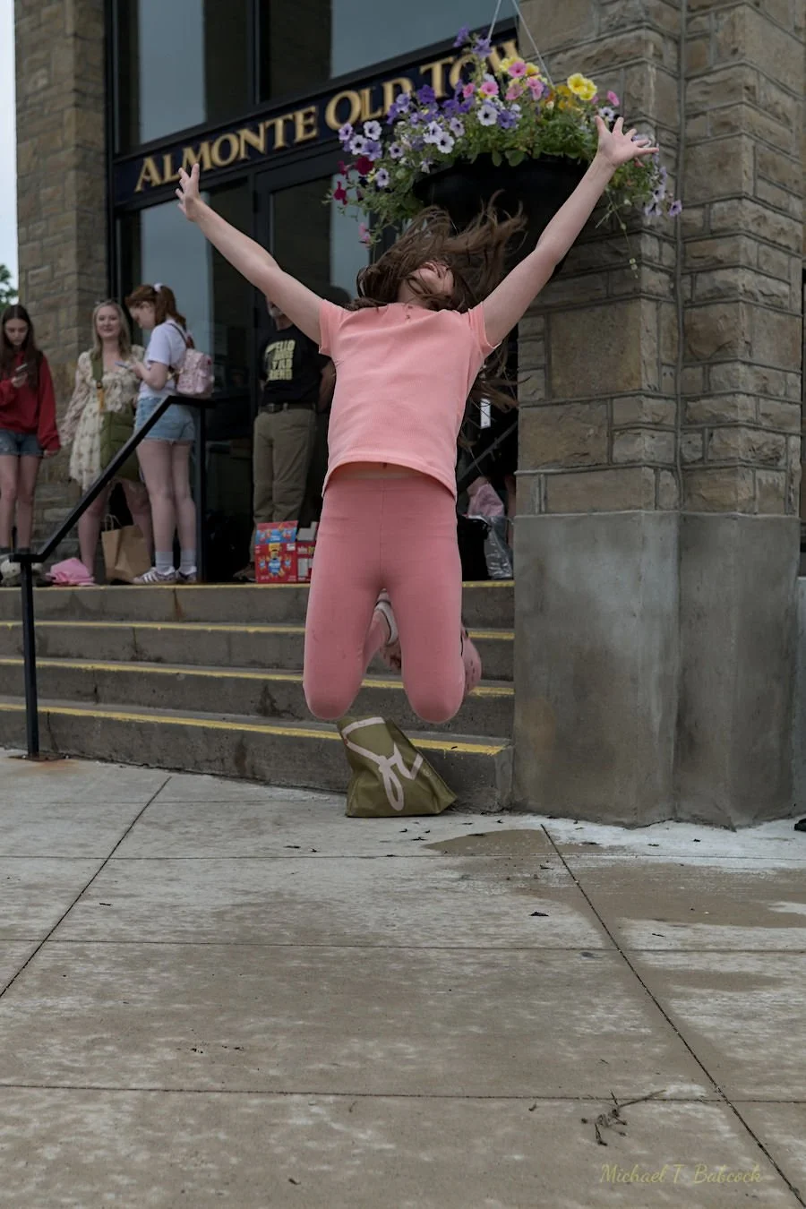 A young woman in a pink outfit is jumping in front of a group of people outside a building with a sign that reads "Al Monte". She has her arms raised and appears joyful. Several girls are standing on the steps in the background, some holding phones. 