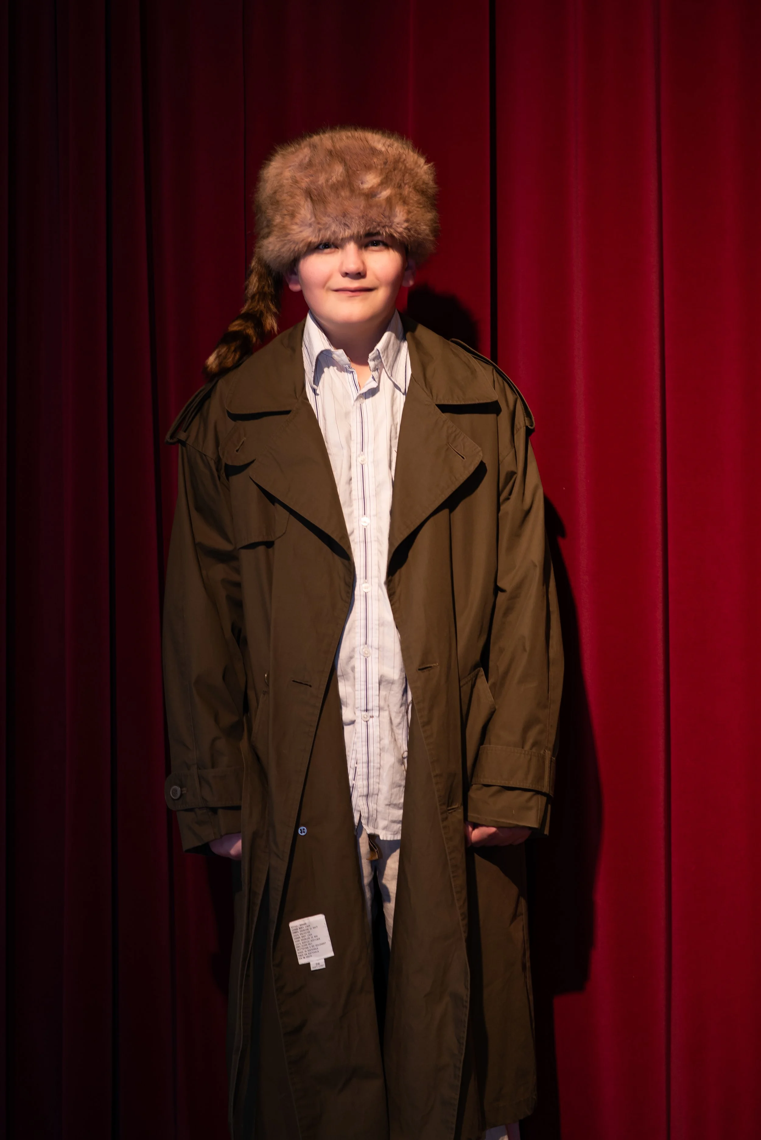 Person wearing a large fur hat and a brown coat standing in front of a red curtain.
