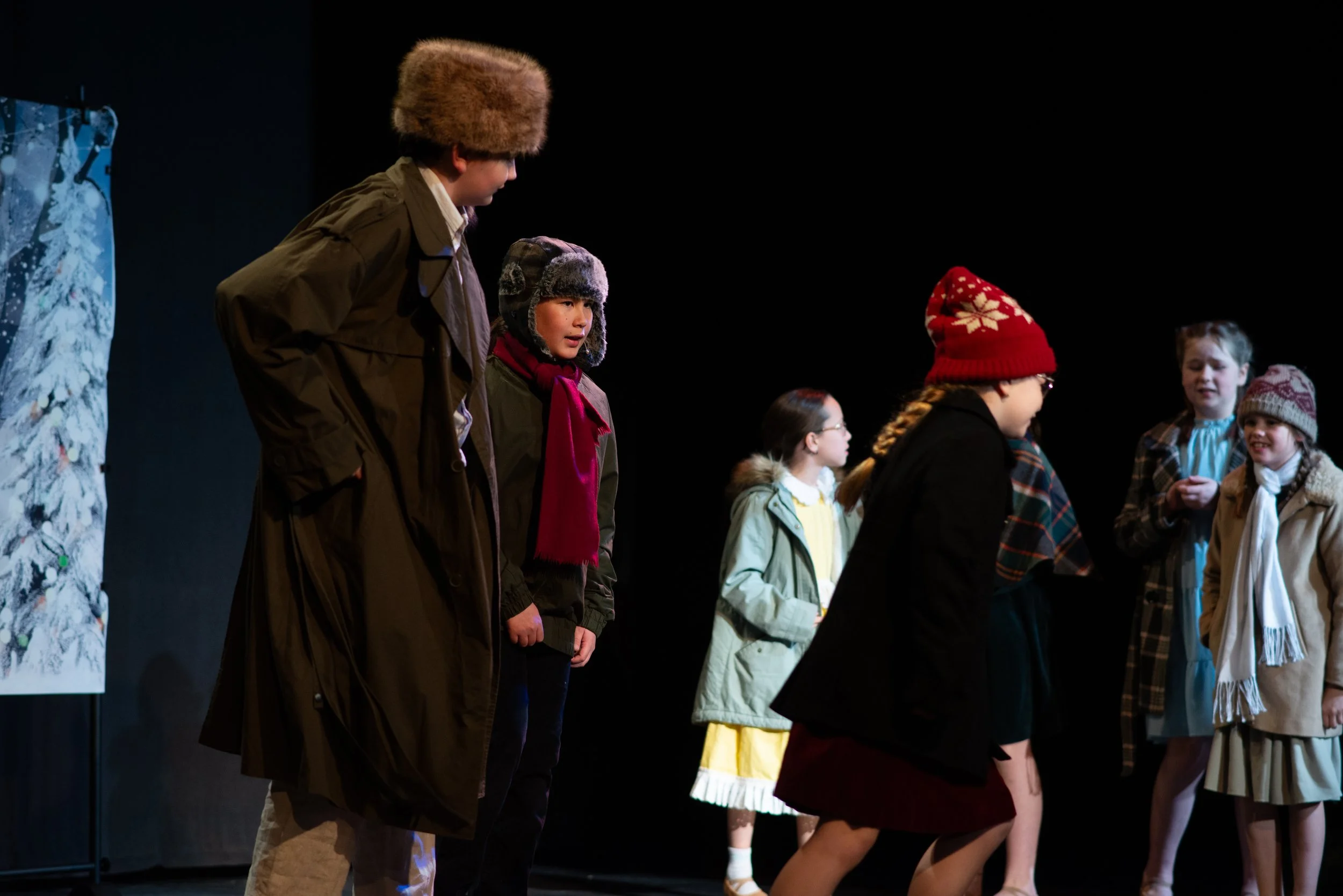 Children dressed in winter clothing standing in line on stage, with a winter-themed backdrop on the left.