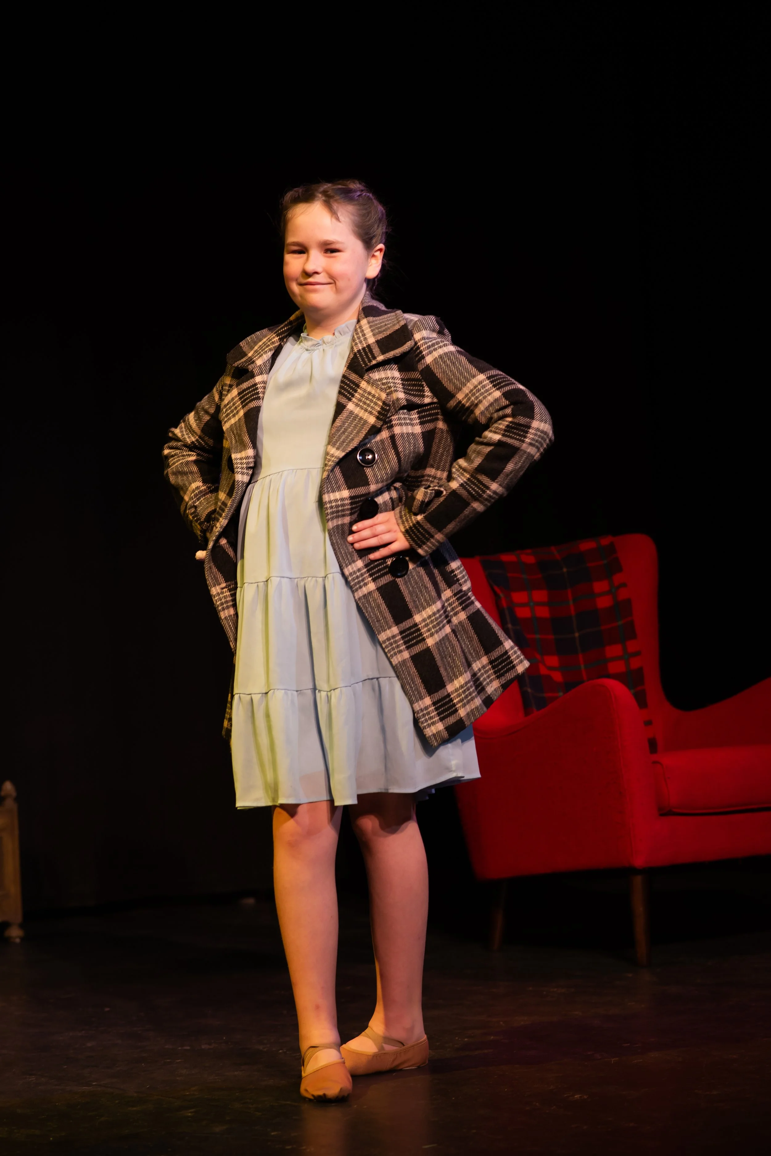 A young girl standing on a stage with hands on her hips, wearing a plaid coat, light-colored dress, and ballet shoes, with a red armchair with a tartan pattern in the background.