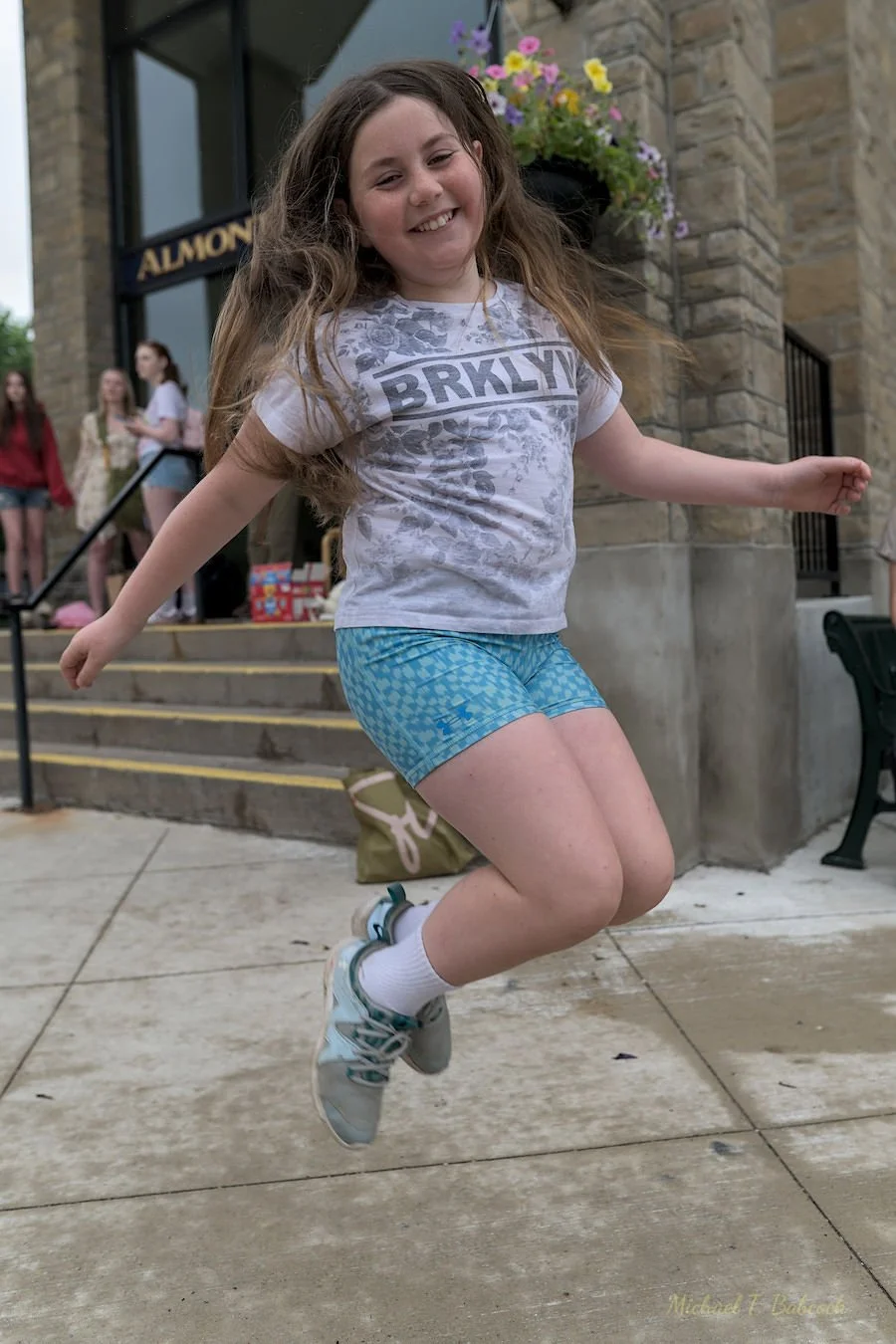 A girl with long, wavy brown hair jumping on a wet sidewalk outside a building with stone walls and stairs, smiling at the camera.