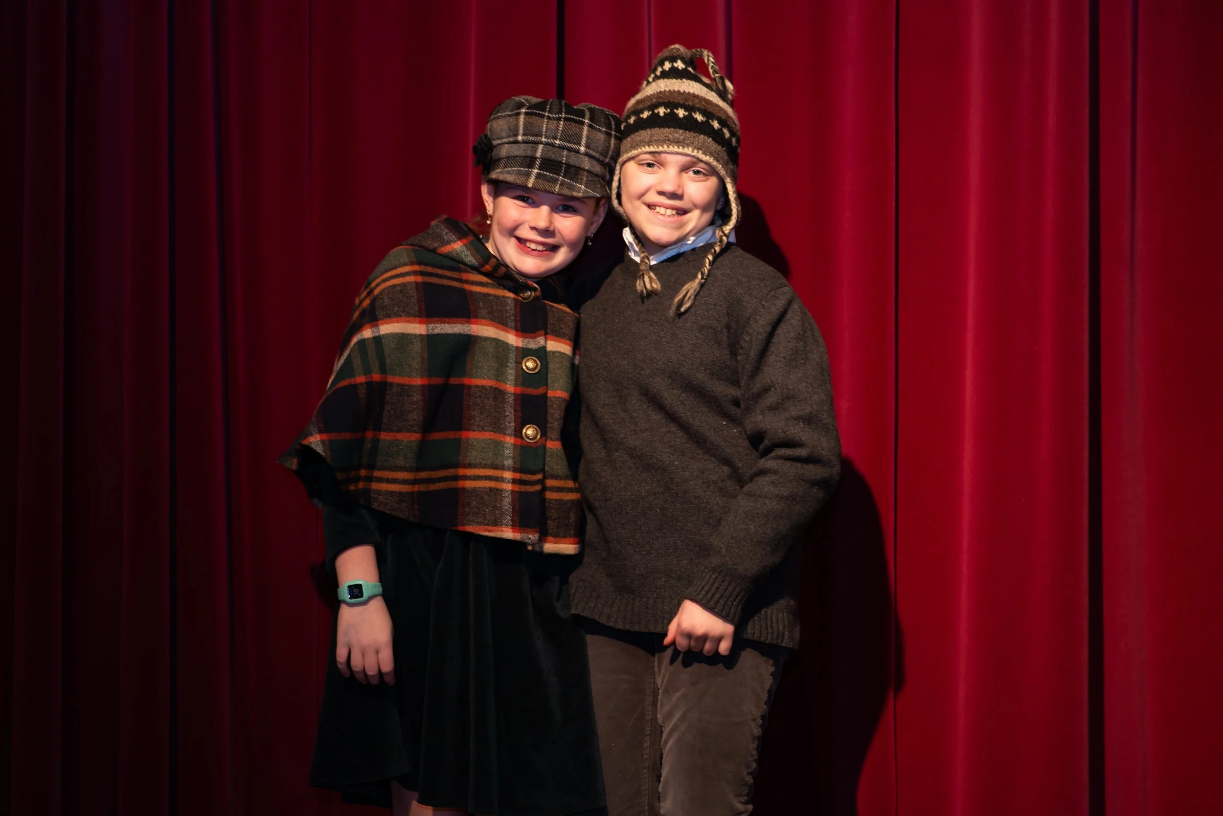 Two children smiling and standing in front of a red curtain. One girl is wearing a plaid hat, a plaid coat, and has a smartwatch on her wrist. The boy is wearing a striped knit hat, a gray sweater, and a collared shirt underneath.