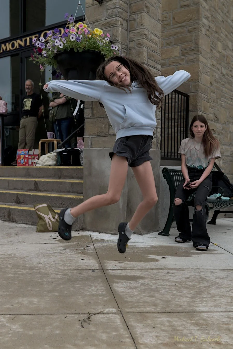 A girl in a white hoodie and black shorts is jumping with her arms spread wide in front of a stone building with a flower basket hanging above her. A girl with long hair and a ripped black pant is sitting on a bench nearby, and a man and a woman are 
