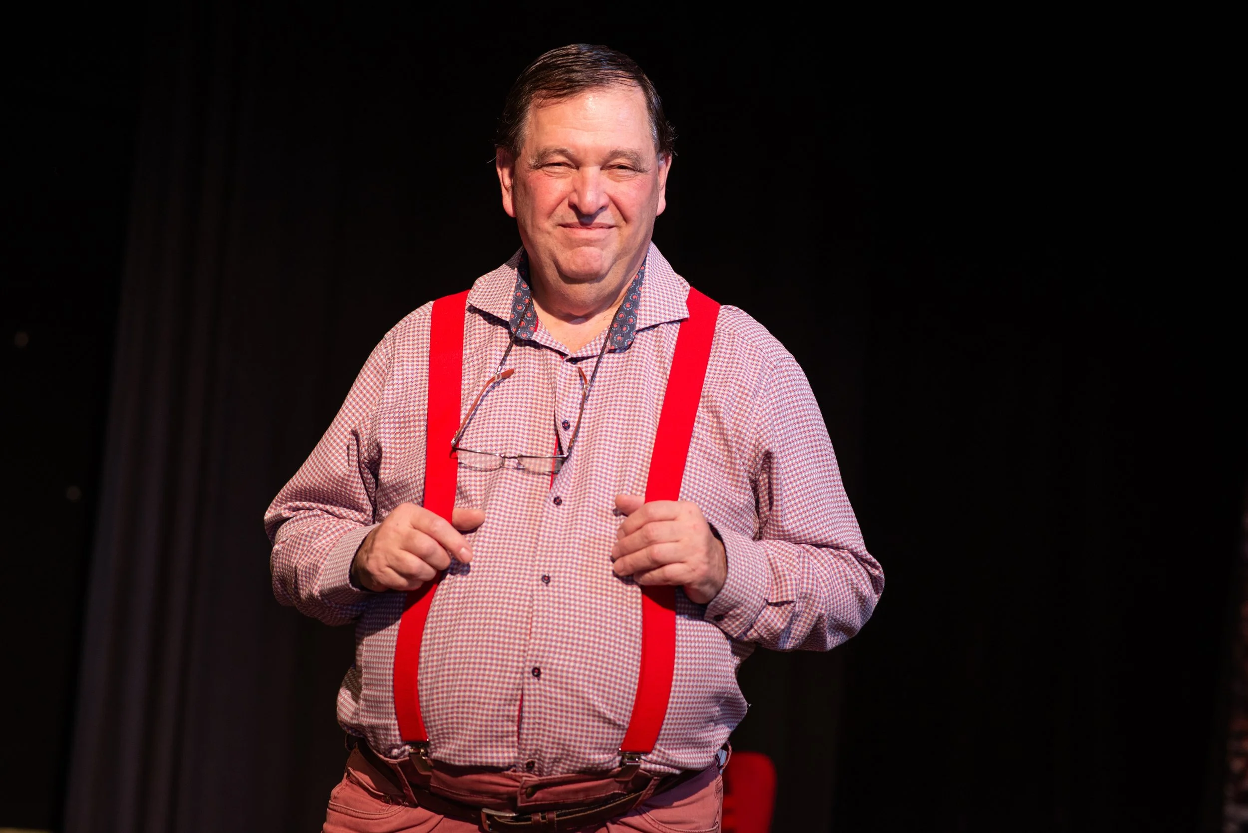 A man on stage wearing a checkered shirt, red suspenders, and glasses around his neck, smiling.