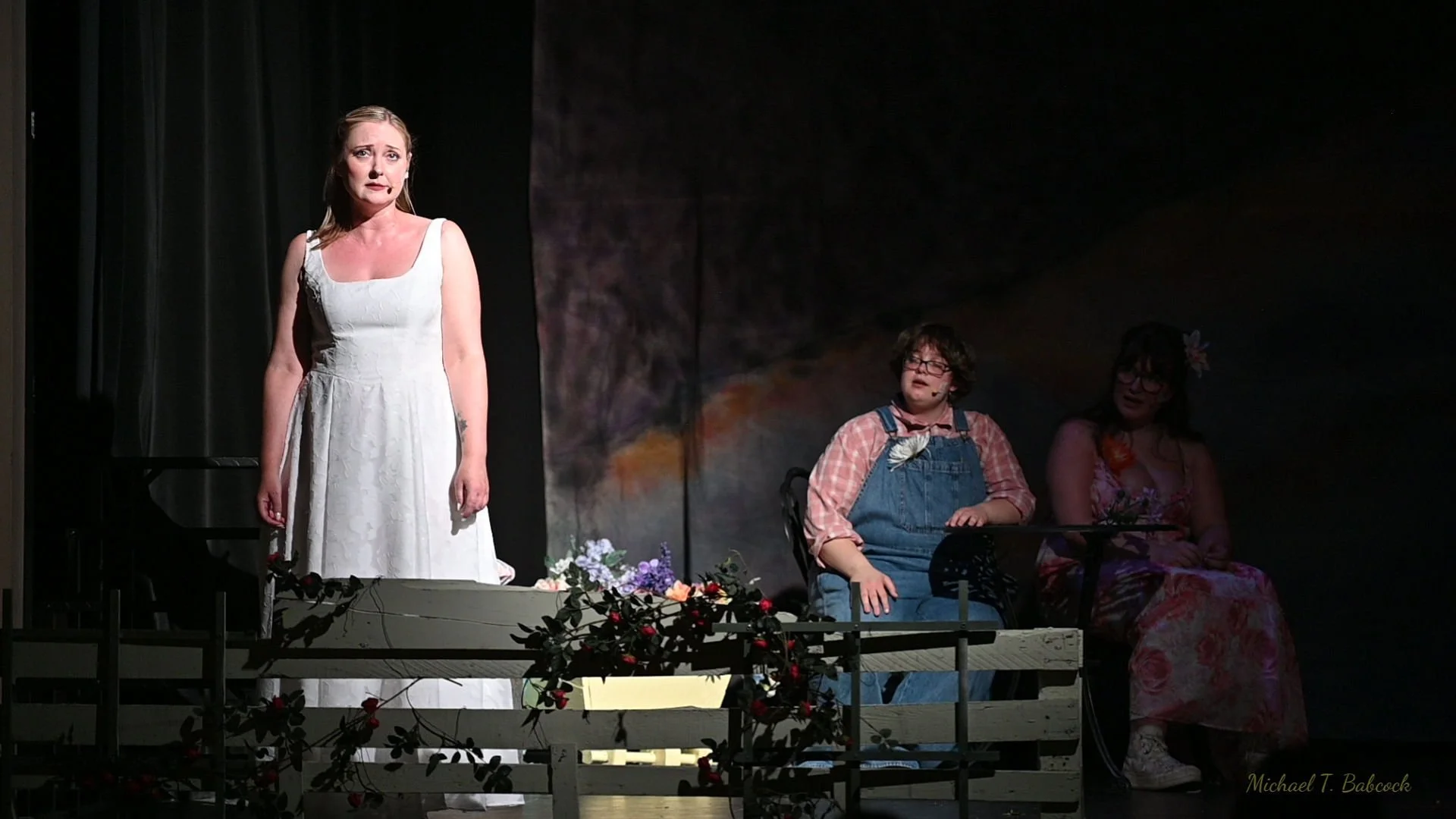 A stage scene from a theatrical performance with female Jesus standing in front, and two seated actors, one in overalls and a checked shirt, and the other in a floral dress. The stage has floral decorations and a dark backdrop.