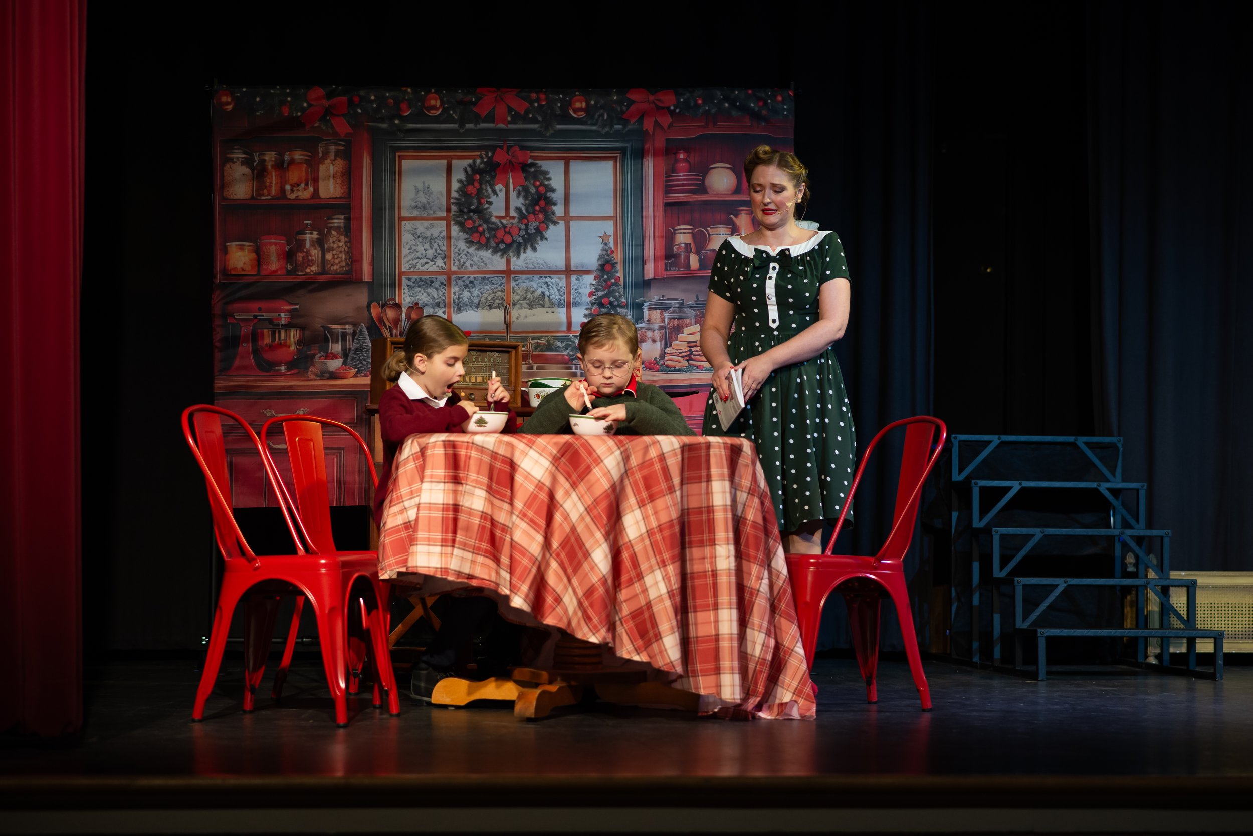 Stage scene of a Christmas play with a backdrop of a kitchen decorated for Christmas, featuring a wreath and window. Two children sit at a table with red chairs, eating from bowls, while a woman in a polka dot dress stands nearby. Red curtains frame 