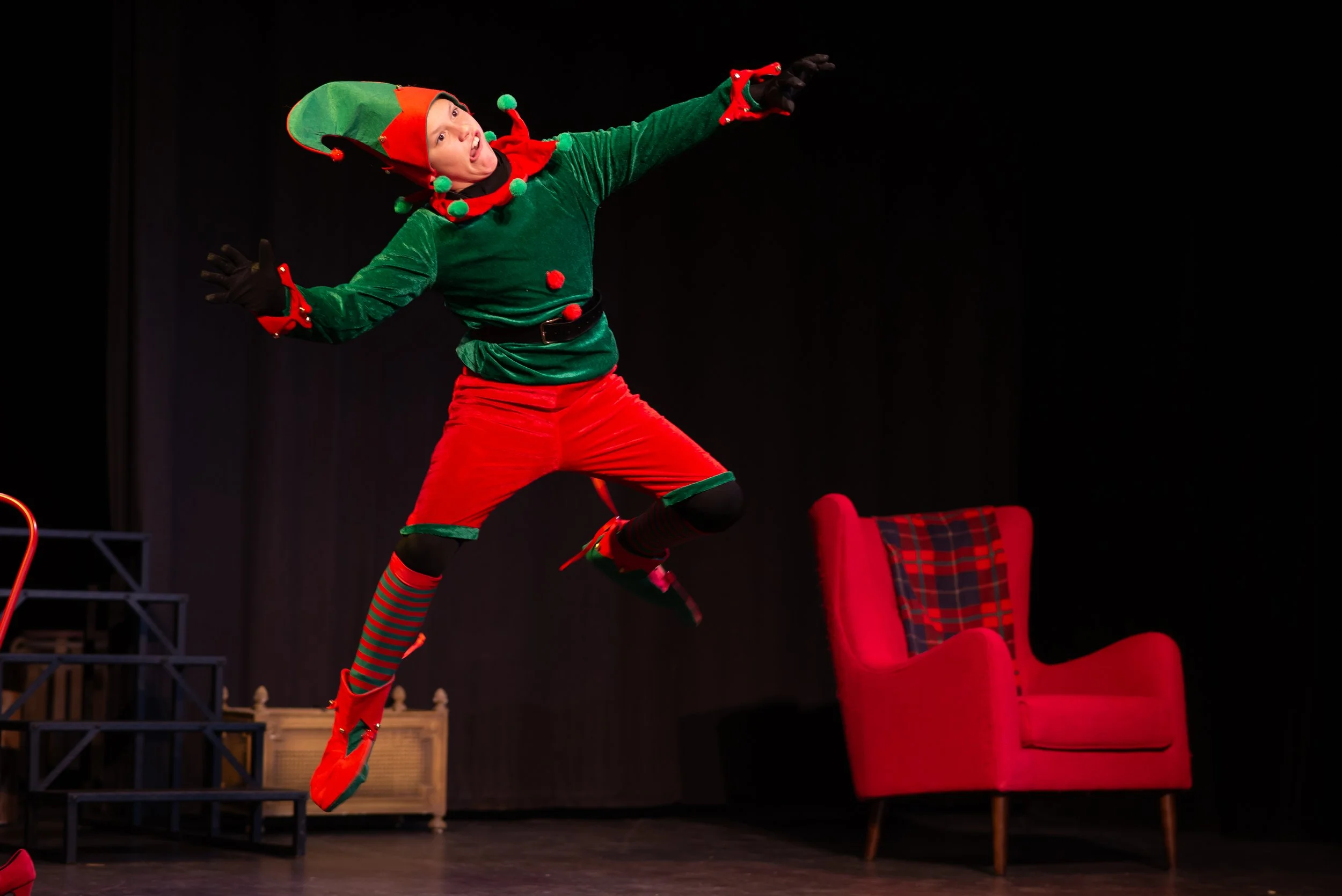 A person dressed as an elf, airborne in a playful pose on stage with red chairs and a red plaid chair in the background.