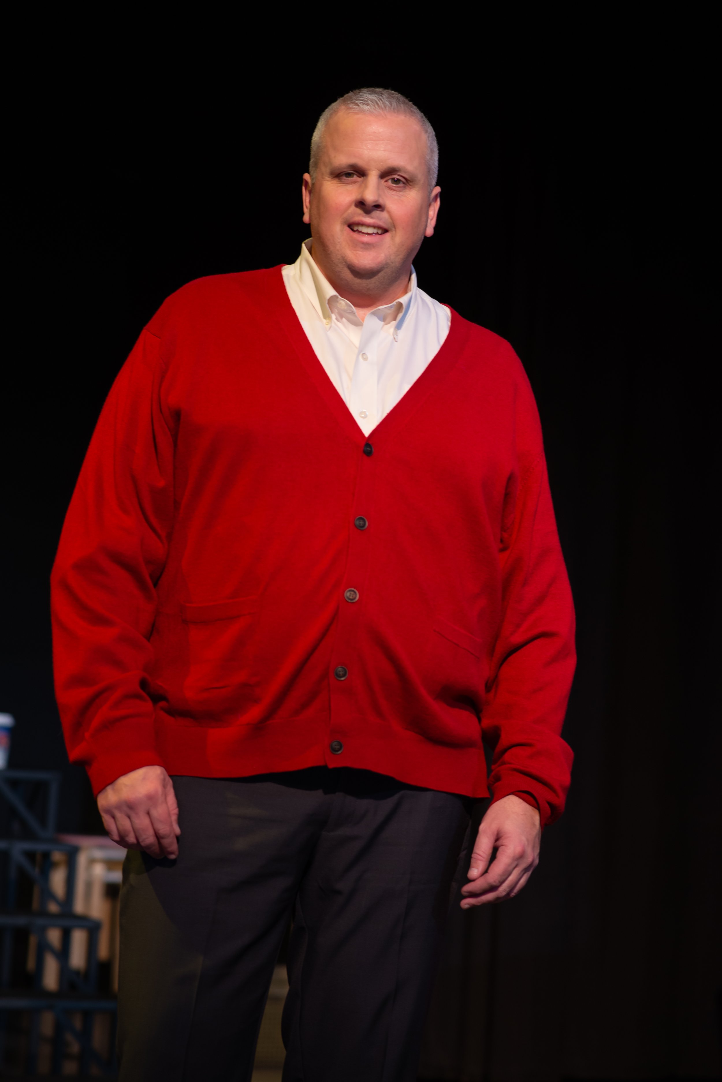 A man with short, gray hair, wearing a white shirt and red cardigan, standing in front of a black background.