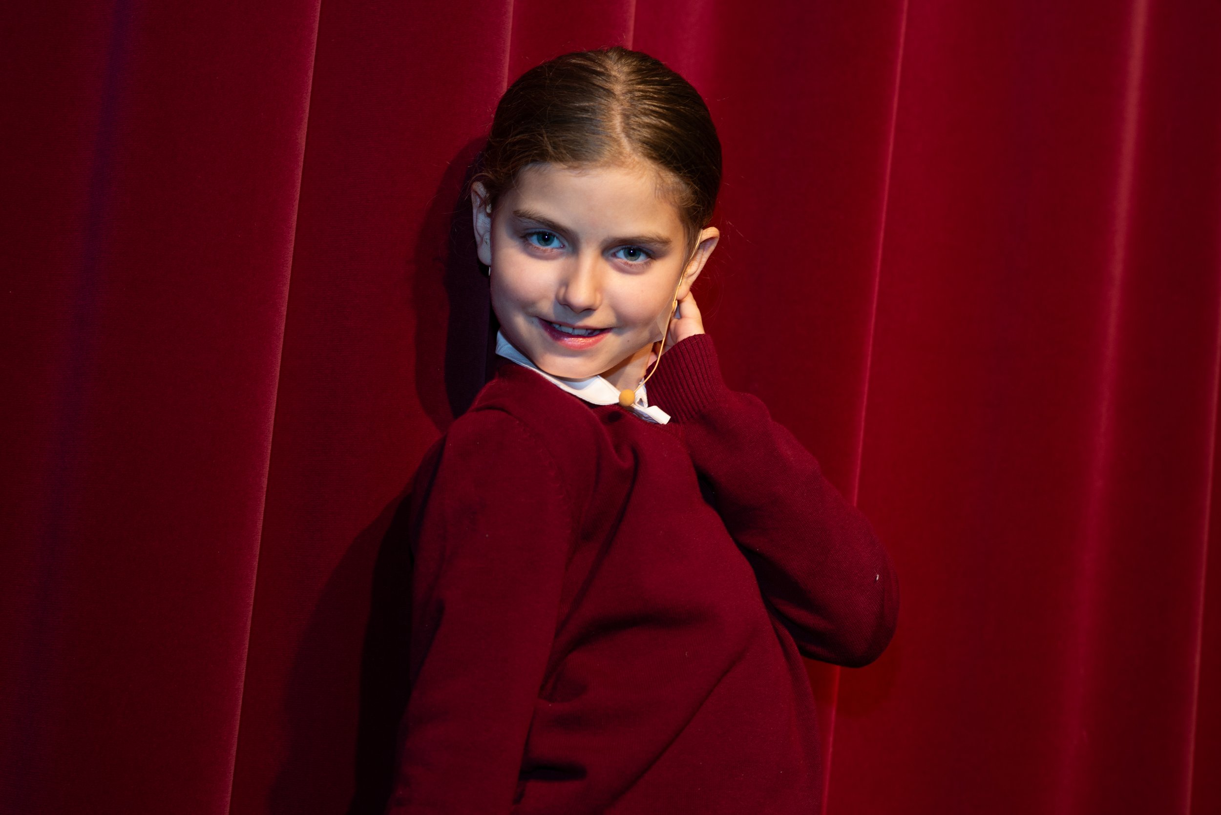 A young girl with long brown hair and blue eyes, wearing a maroon sweater, sitting against red velvet curtains, smiling and touching her ear.