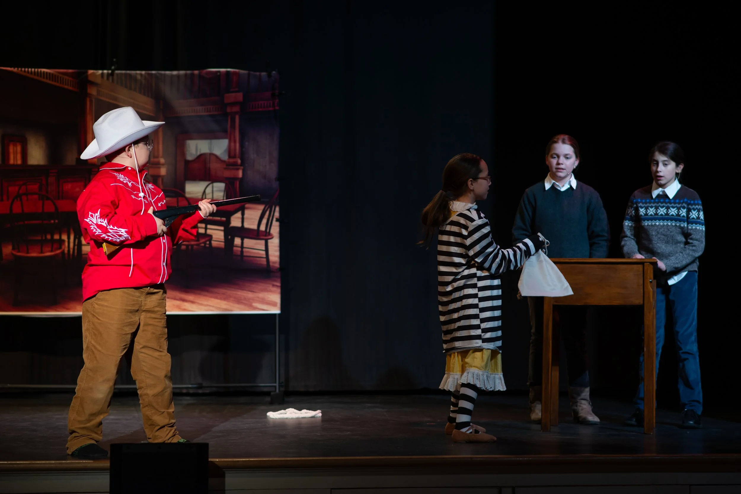 Children performing a play on stage, with one girl in striped dress and three others watching, while a boy in cowboy attire holds a rifle, set against a backdrop resembling a Western town.
