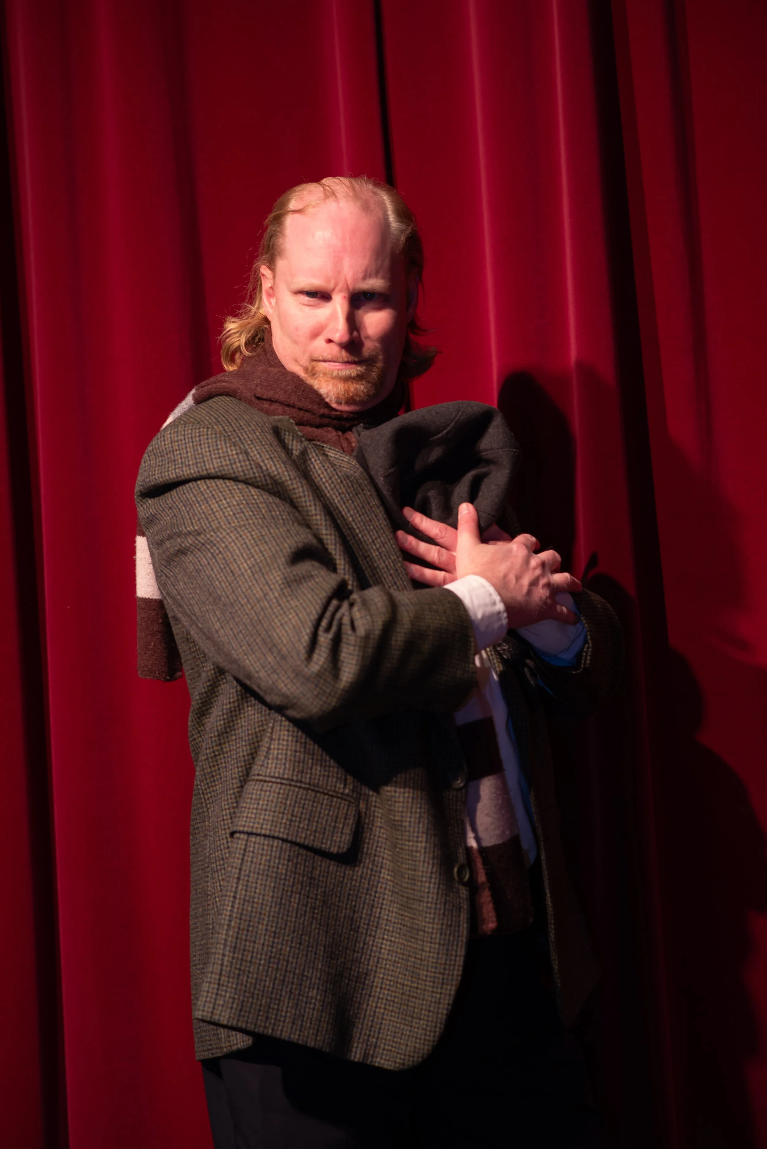 A man with long hair and a beard stands in front of red curtains, looking intensely at the camera, holding a jacket close to his chest with both hands.