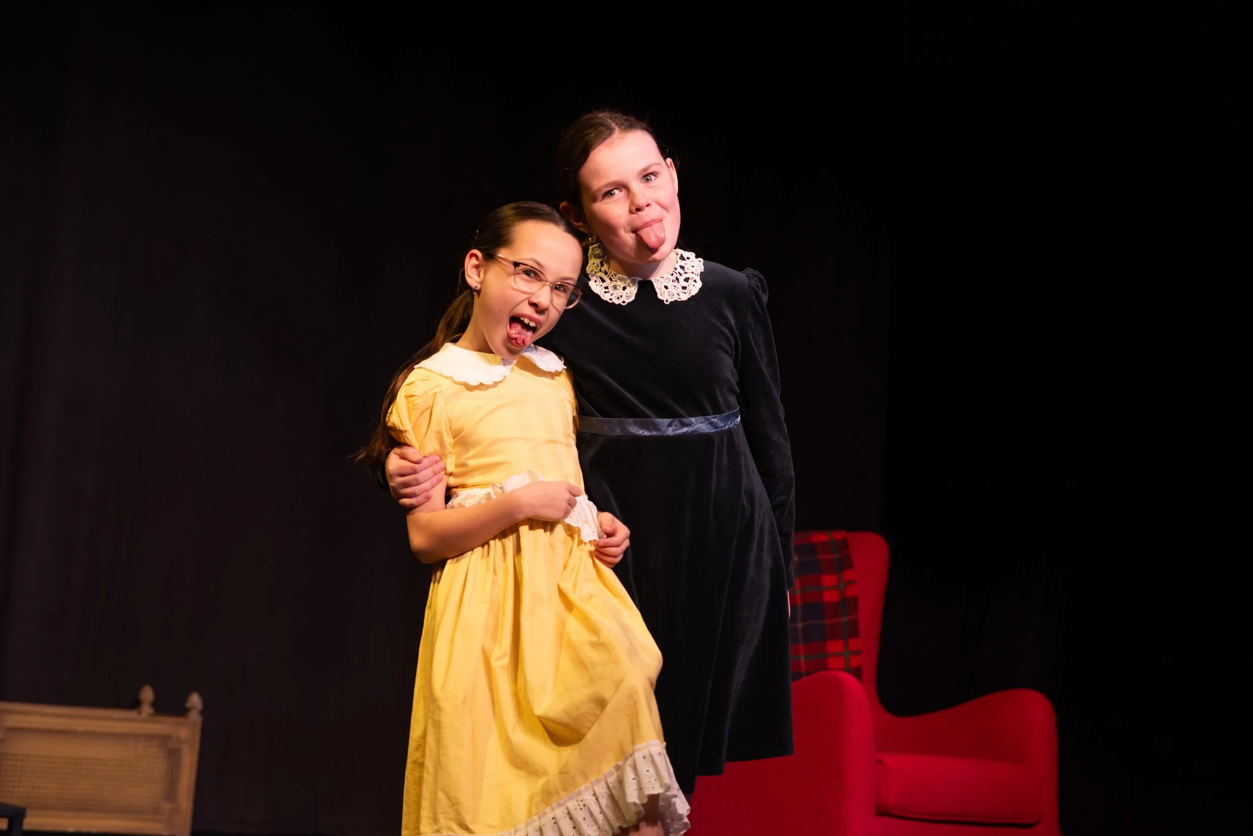 Two girls on stage, one in a yellow dress making a silly face with tongue out, the other in a black dress with a lace collar making a silly face with tongue out, standing close together.