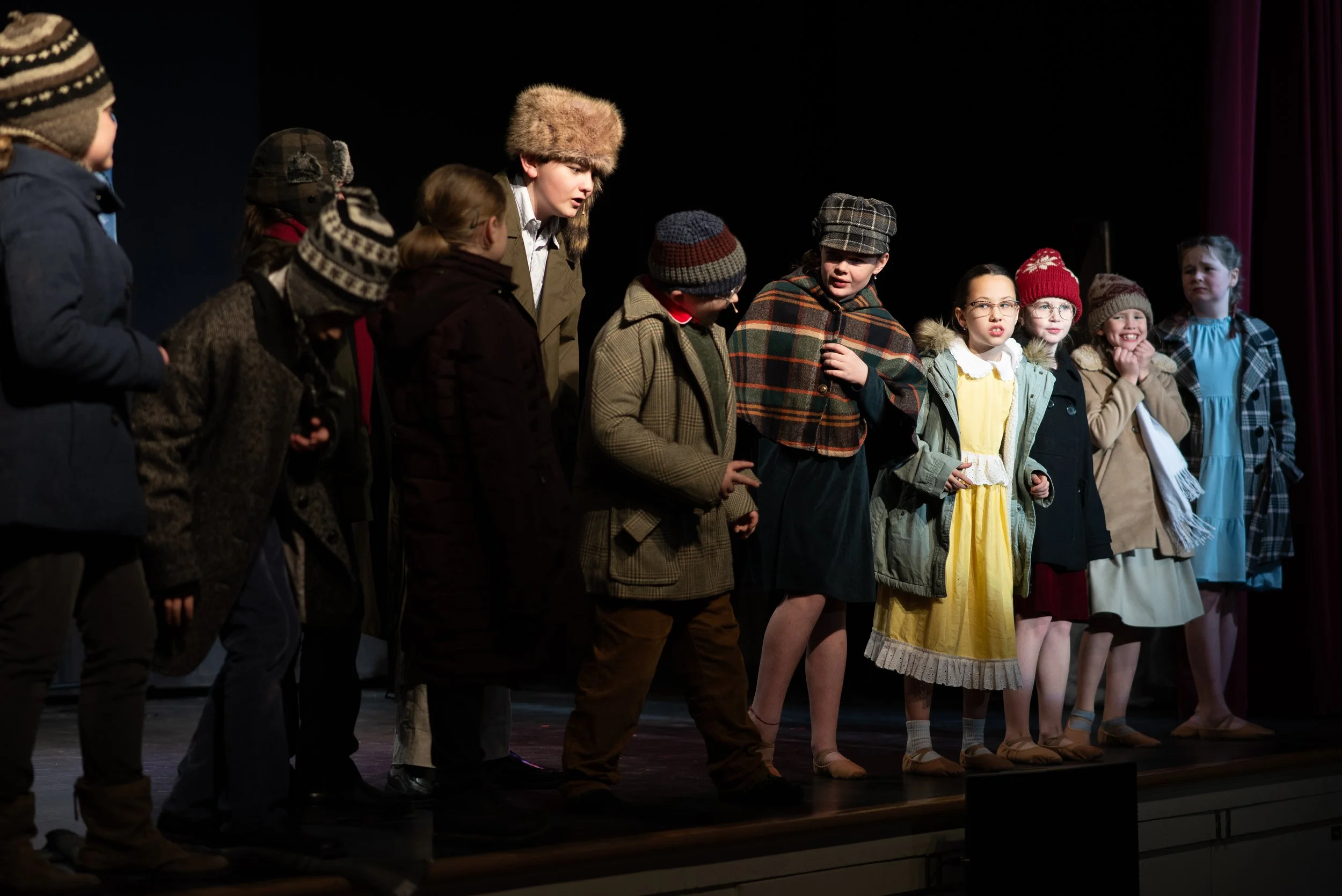 Young children dressed in vintage winter clothing, standing in a line on stage, possibly performing in a play or participating in a school event.