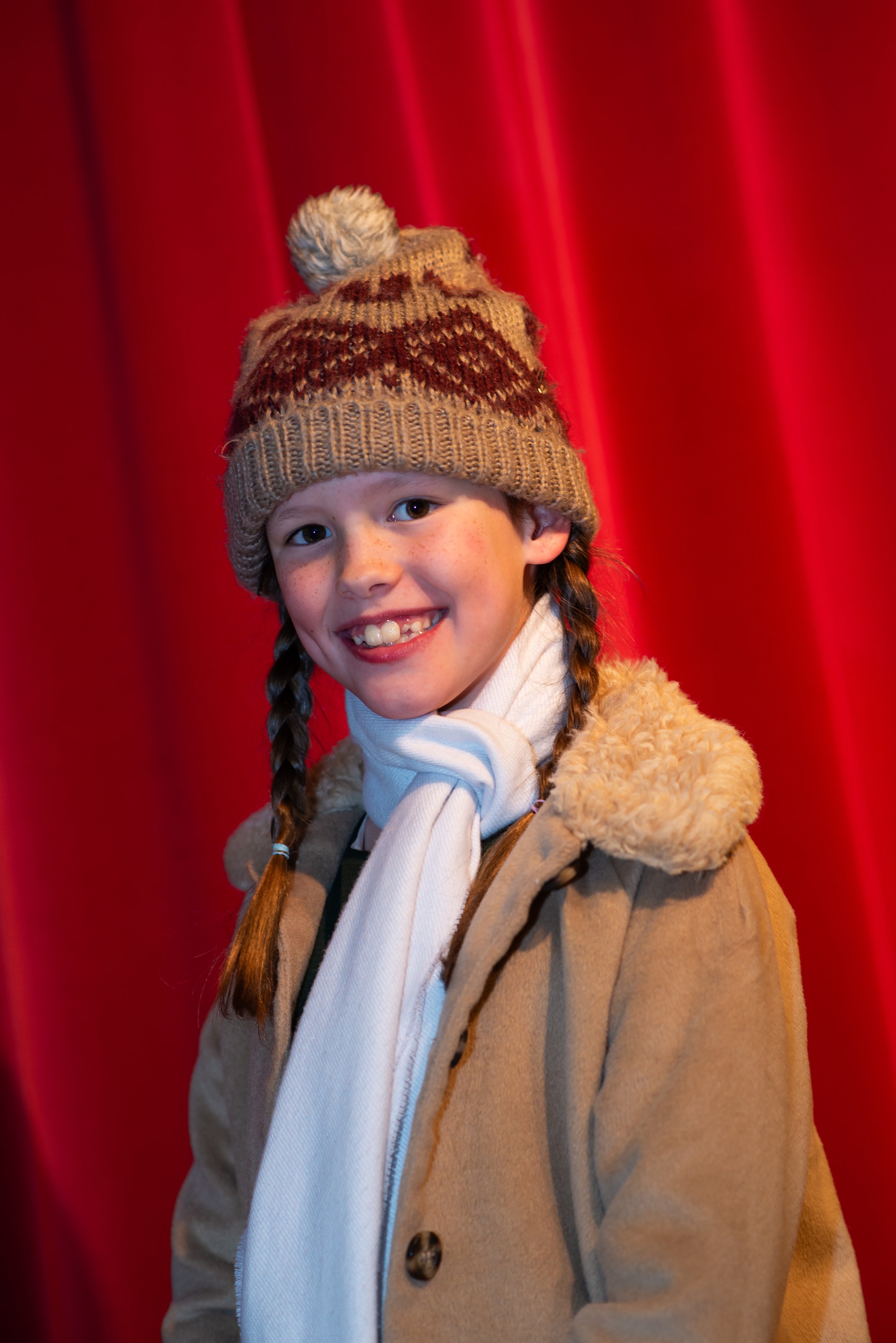 A young girl with braids, wearing a tan coat with faux fur trim and a knitted winter hat with a pom-pom, smiling in front of a red curtain.